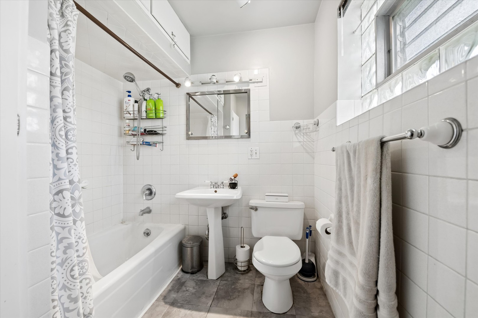 Tiled bathroom with a bathtub, patterned shower curtain, pedestal sink, toilet, and glass-block windows.