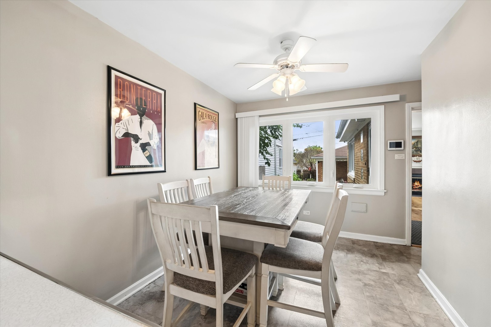Dining room with a gray wood table, white chairs, framed wine posters, and a wide window.