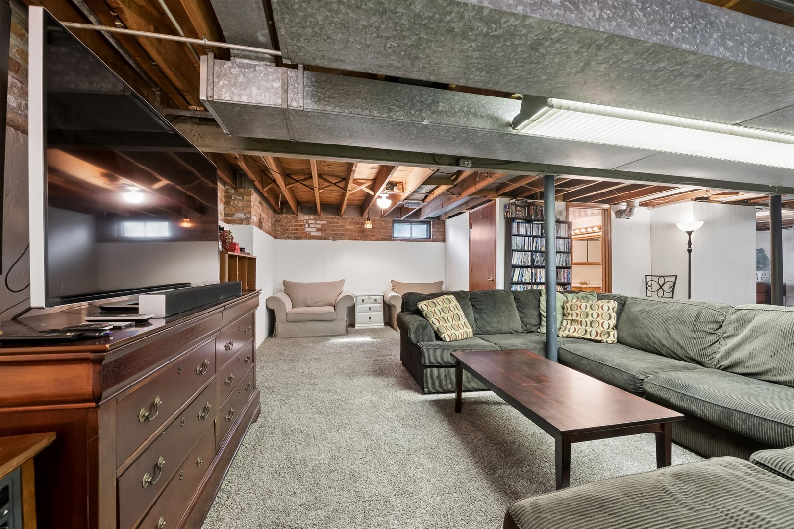 Finished basement family room with a gray sectional, television, exposed beams, and metal ceiling ductwork.