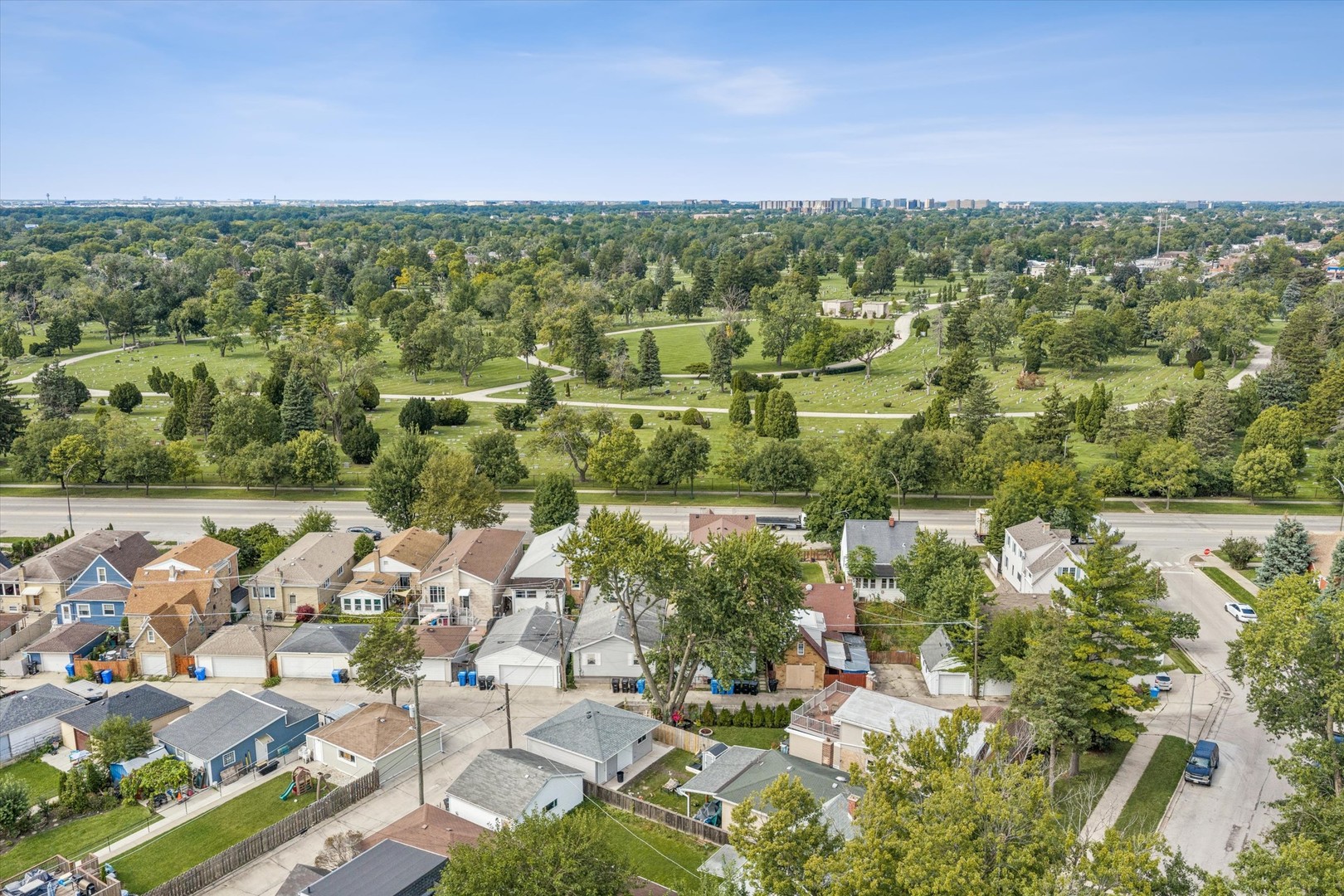 Aerial view of neighborhood homes beside a sprawling, tree-filled cemetery with winding paths.