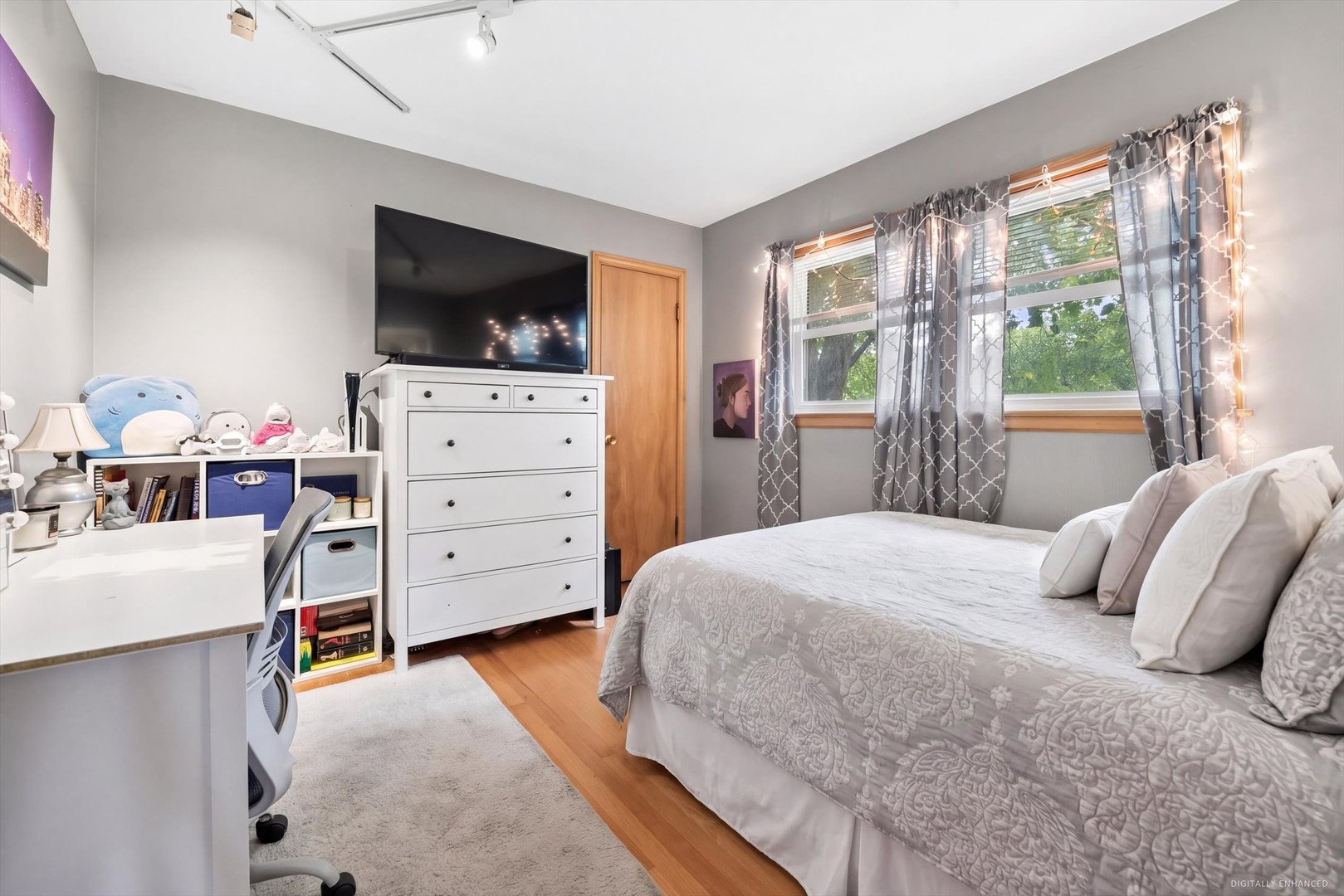 Furnished bedroom with a large bed, television on a white dresser, and two curtained windows.