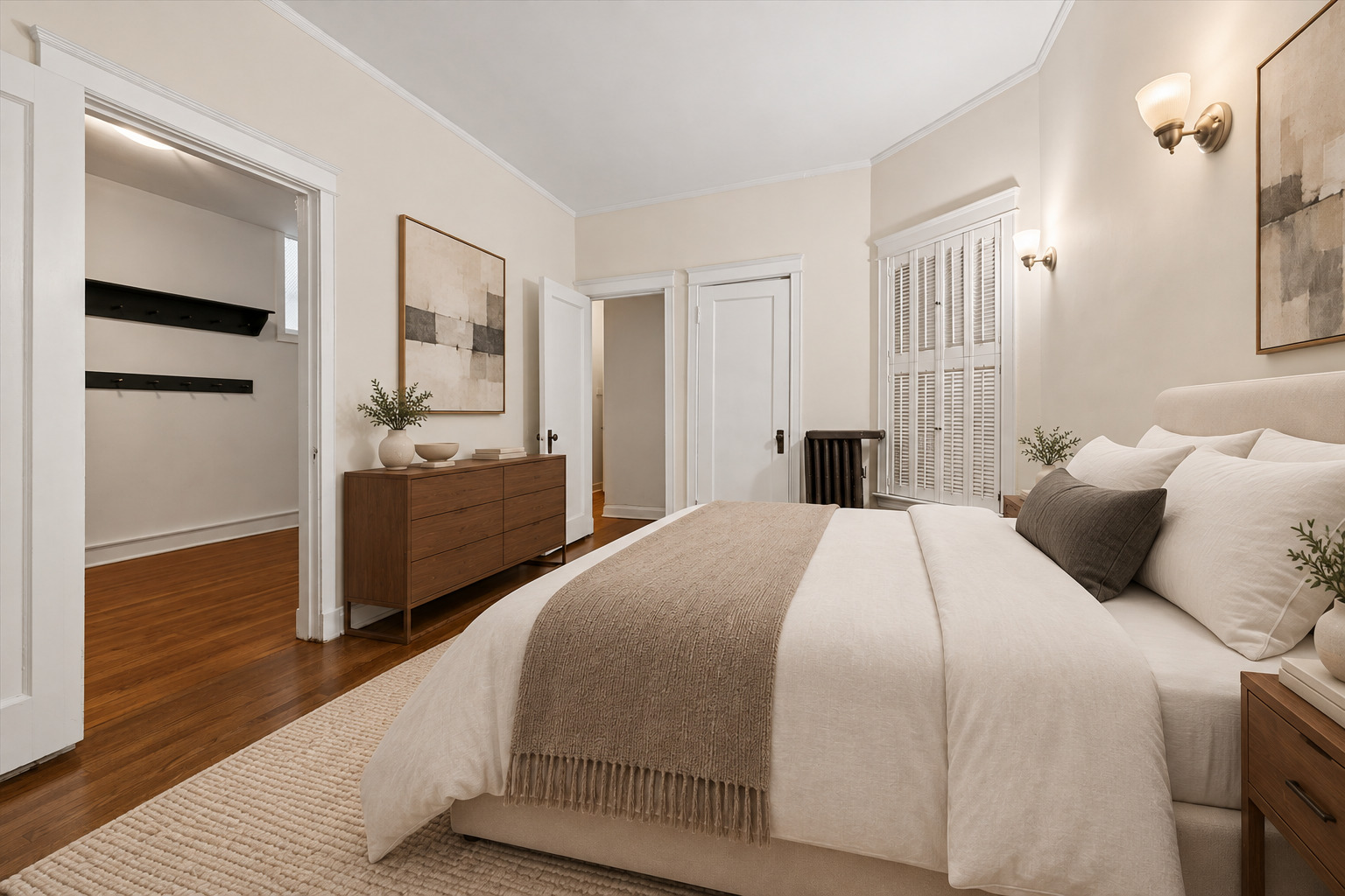 Staged bedroom with a white bed, taupe throw, wooden dresser, and shuttered window.