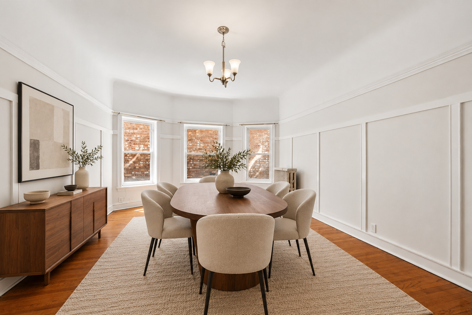 Dining room with an oval wood table, upholstered chairs, white paneled walls, and hardwood floors.