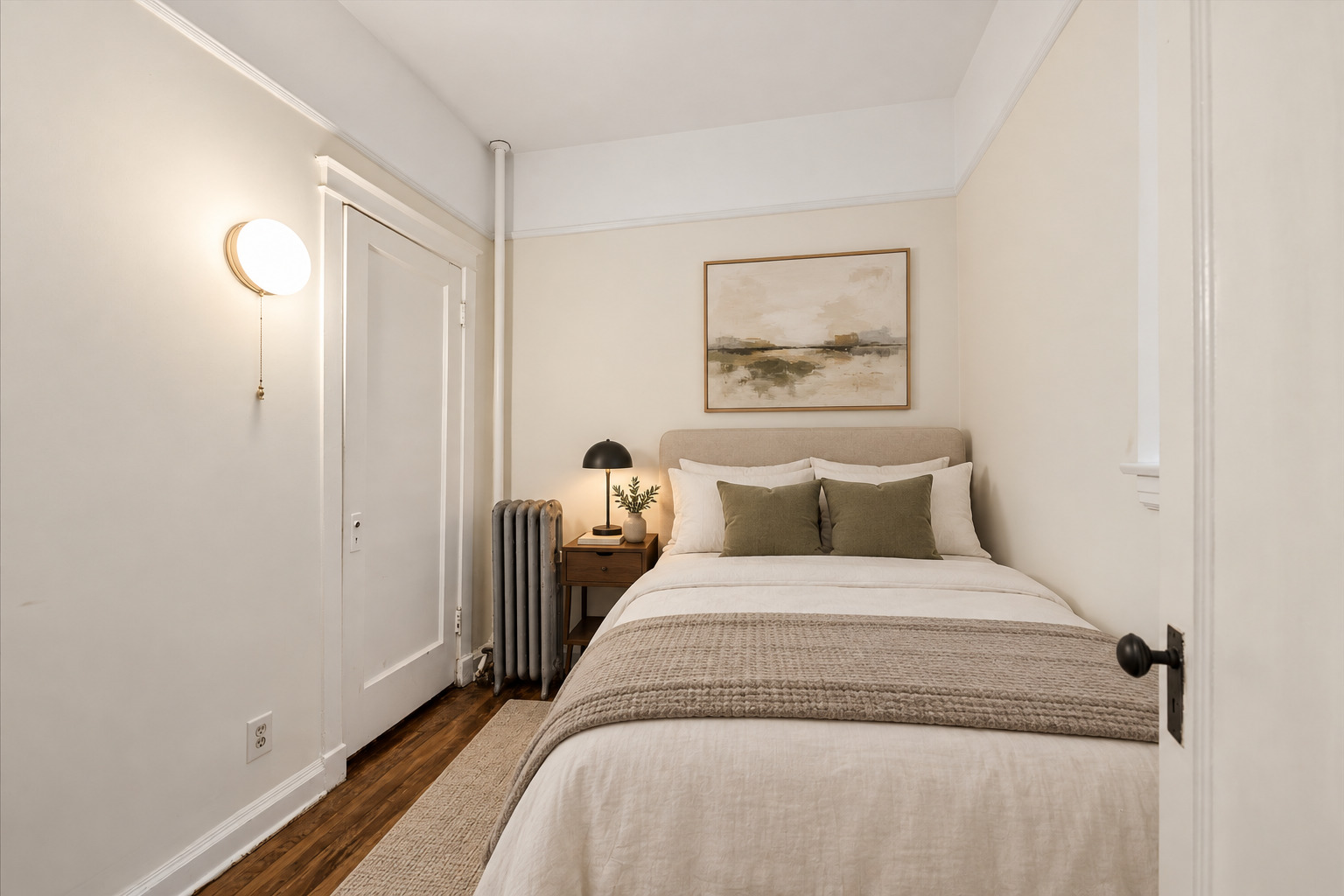 Staged bedroom with a white bed, olive-green pillows, abstract art, and a radiator beside the doorway.