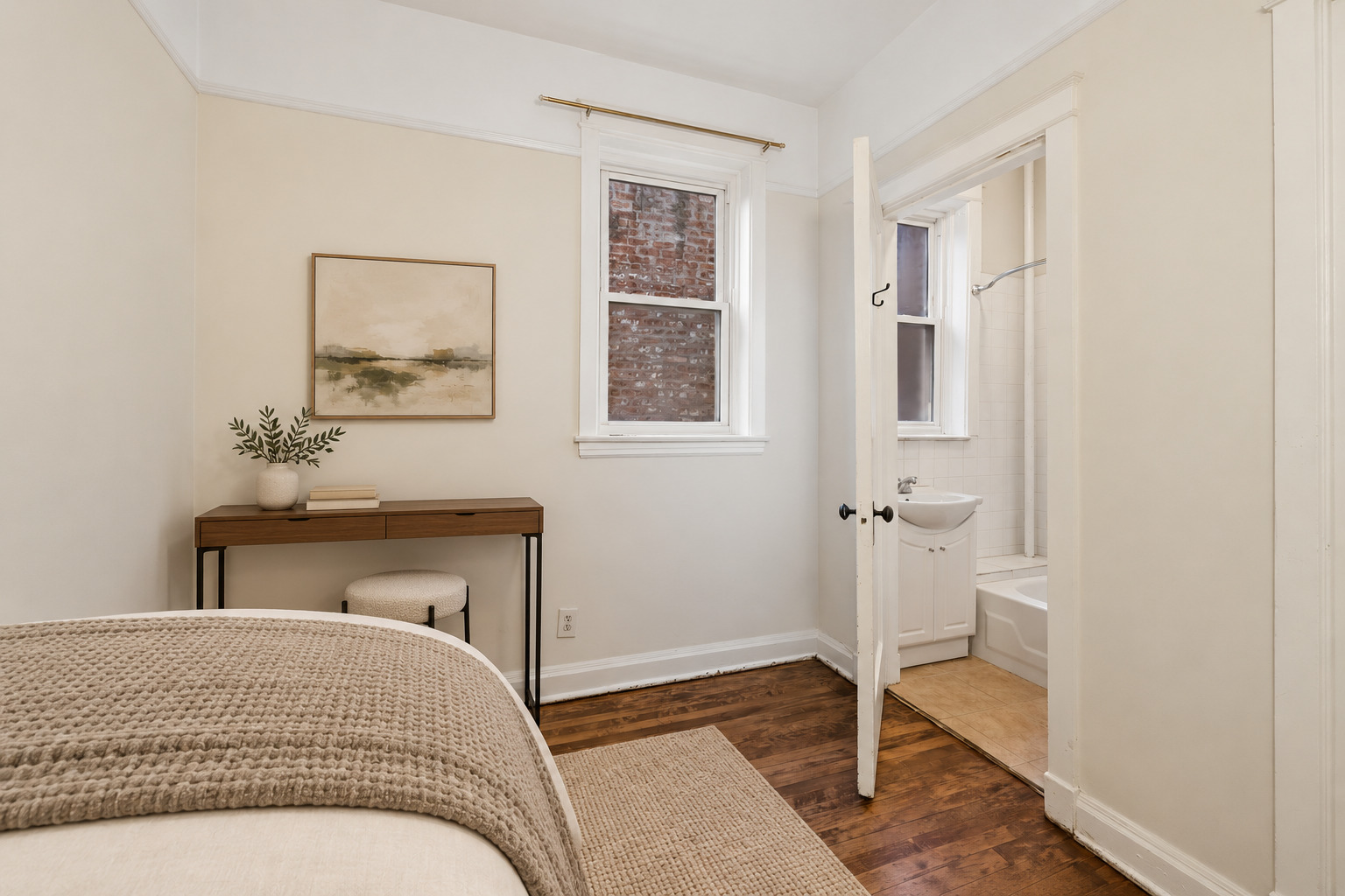 Staged bedroom with a textured cream bedspread, slim desk, dark hardwood floors, and an adjoining bathroom.