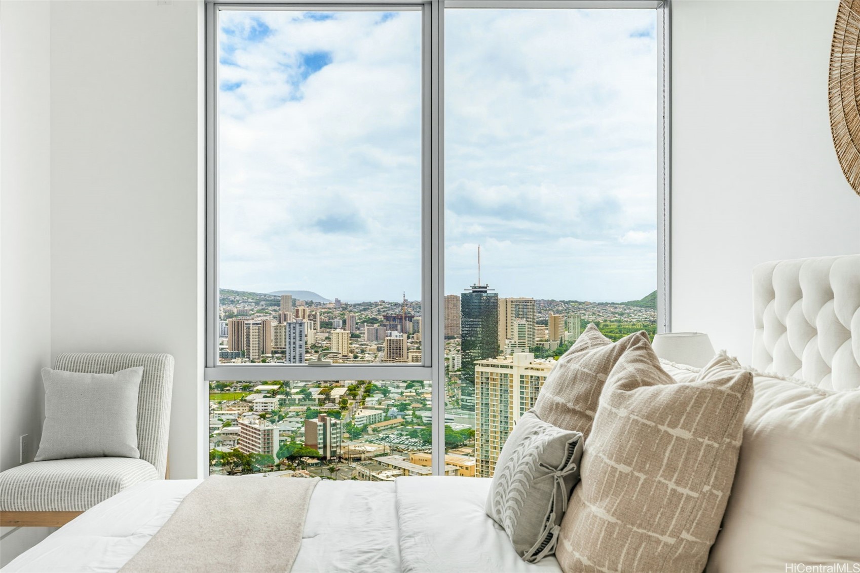 Bedroom with a tufted bed and chair overlooking Honolulu’s skyline through tall windows.