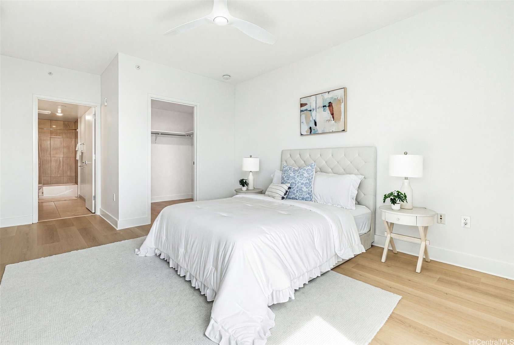 Staged bedroom with white tufted bed, light wood floors, and open bathroom doorway.