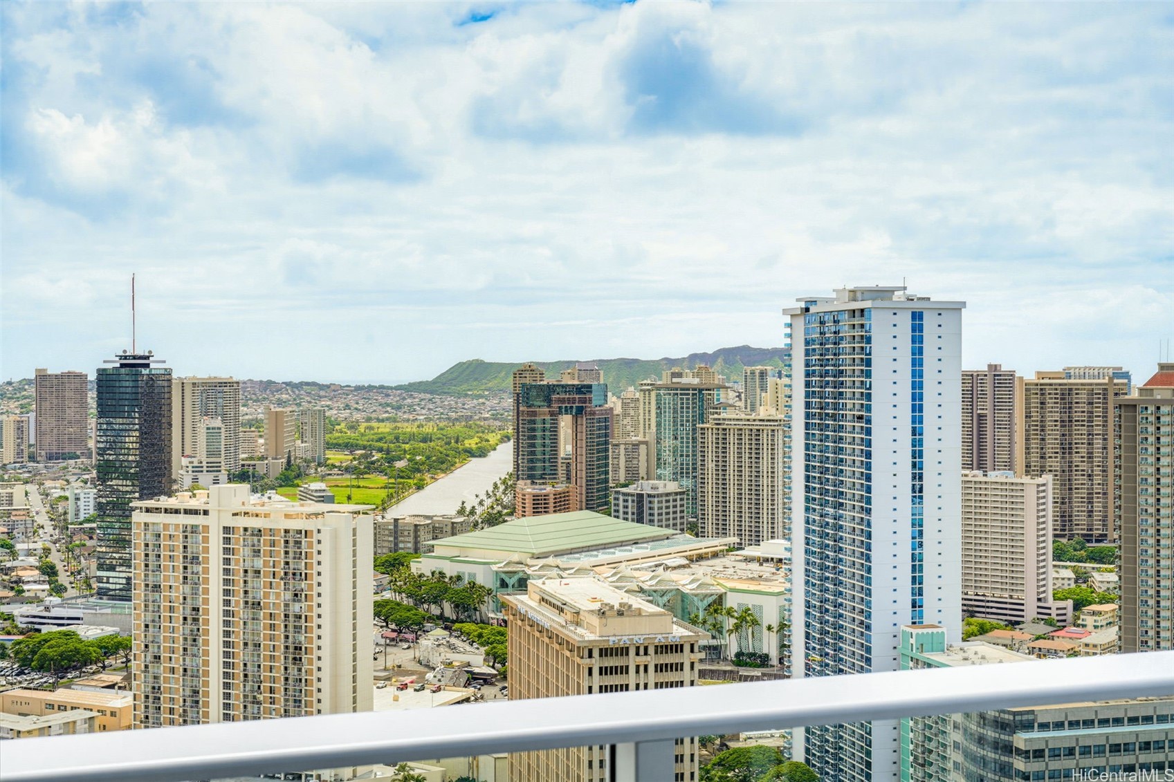 Honolulu city skyline and Ala Wai canal framed by mountains, viewed over a high-rise balcony railing.