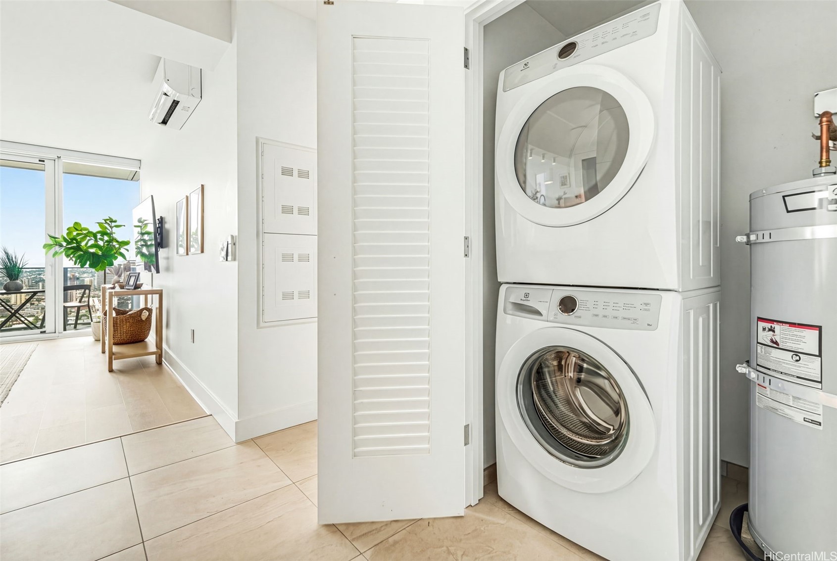 Stacked washer and dryer beside a bright living room with sliding glass doors and a city view.