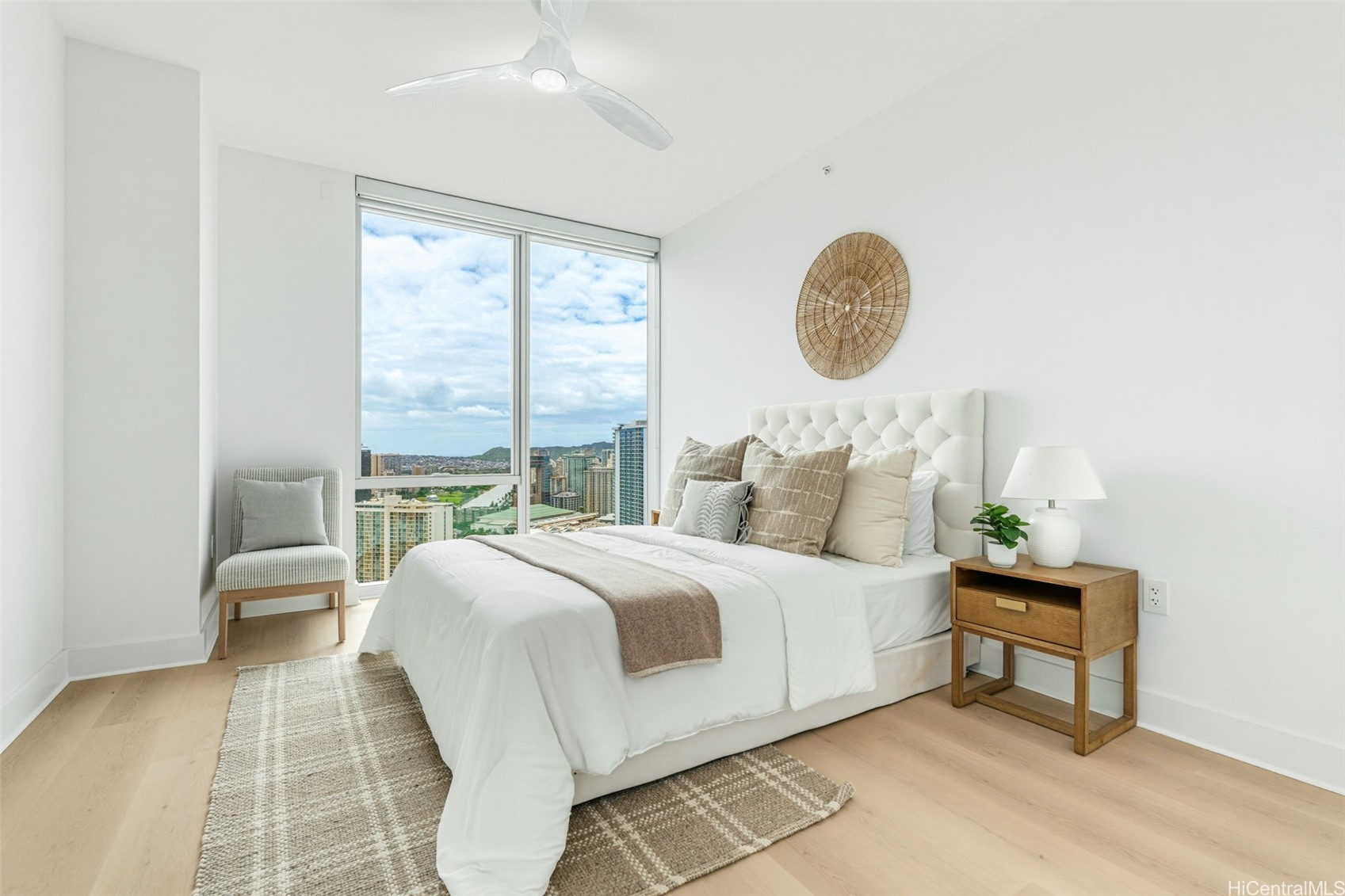 Staged bedroom with a white tufted bed and city views through large windows.
