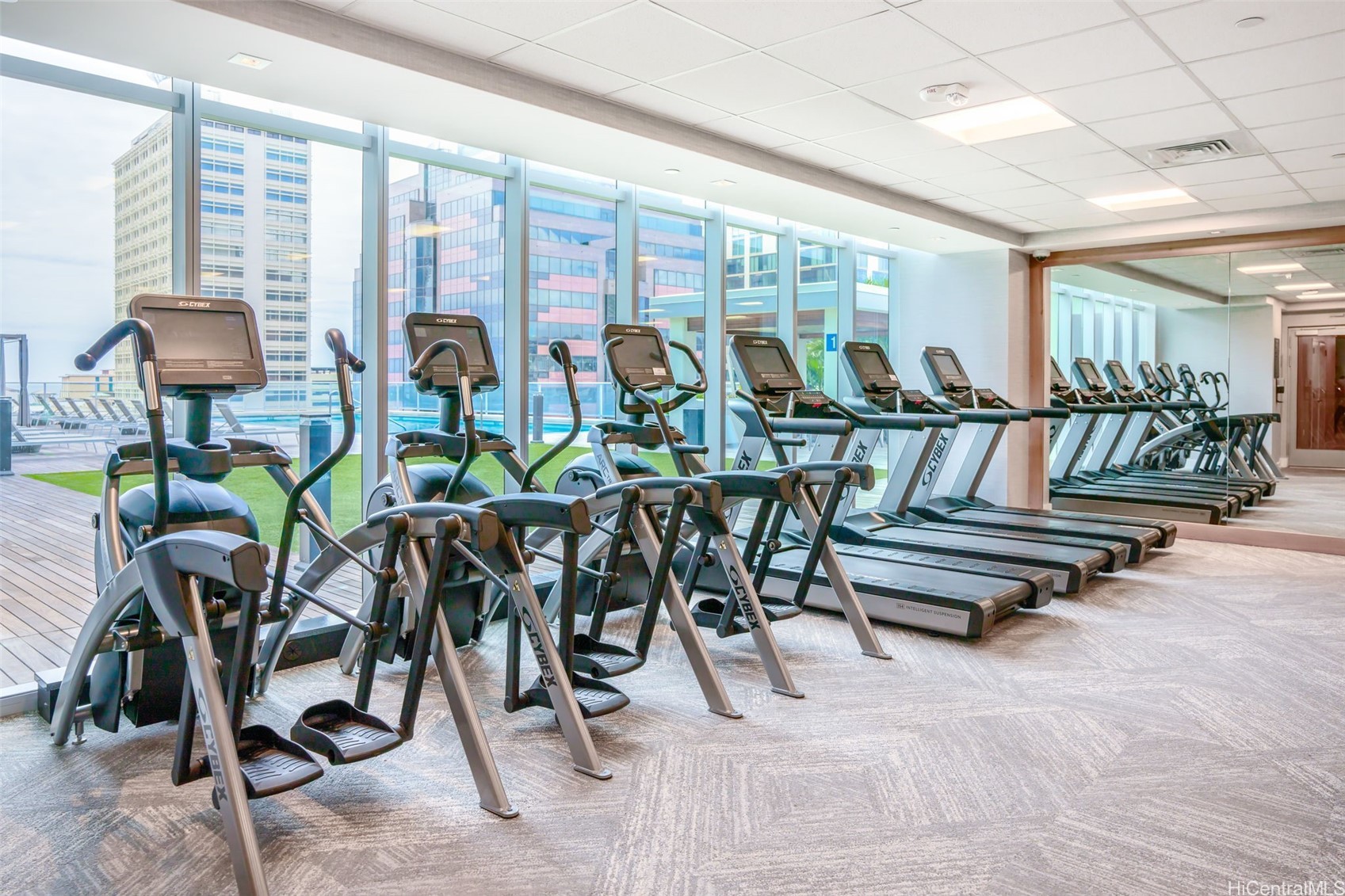 Condominium gym with rows of cardio machines beside floor-to-ceiling windows overlooking nearby buildings.