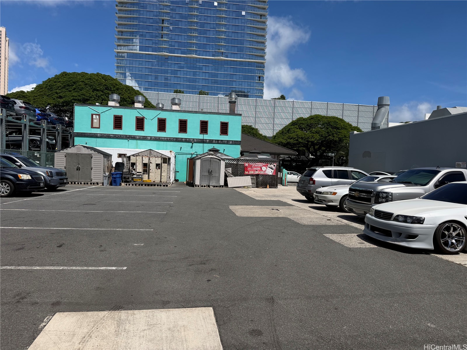 Parking lot beside a turquoise building, with parked cars and a glass high-rise behind it.