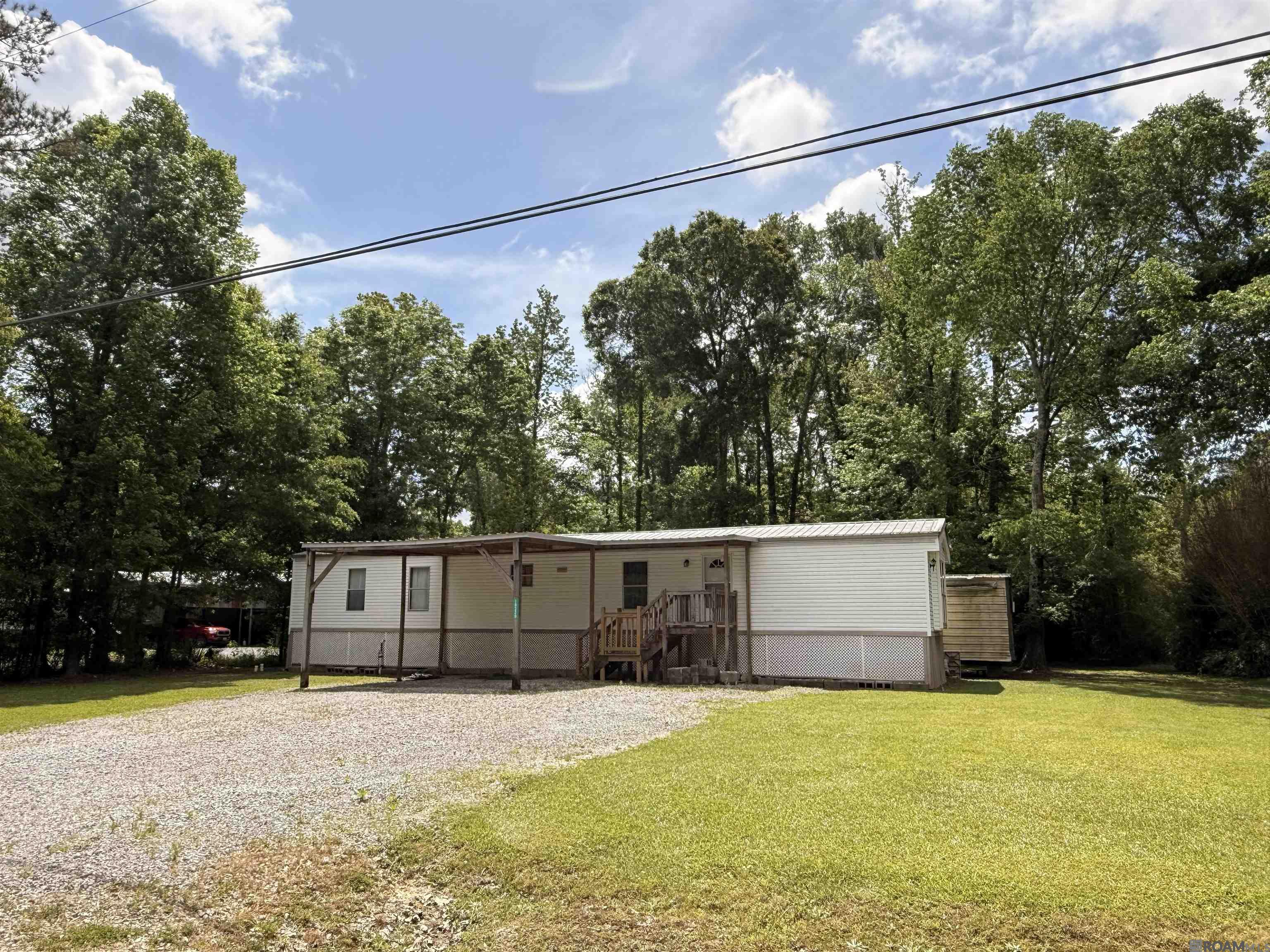 Manufactured home with a covered porch, gravel driveway, and mature trees.