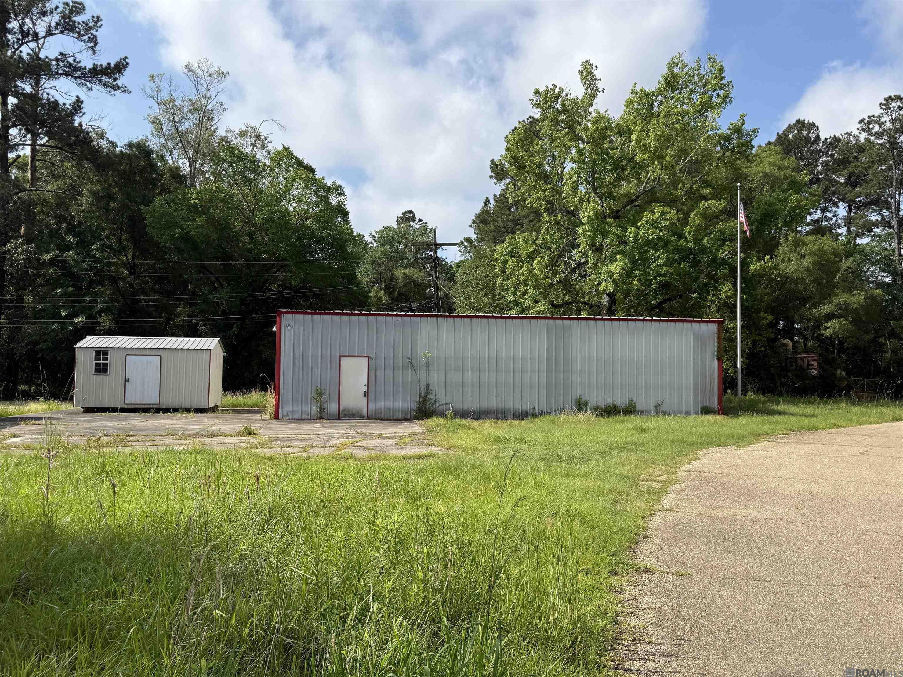 Long metal-sided building with red trim beside a smaller outbuilding, tall grass, and a paved drive.