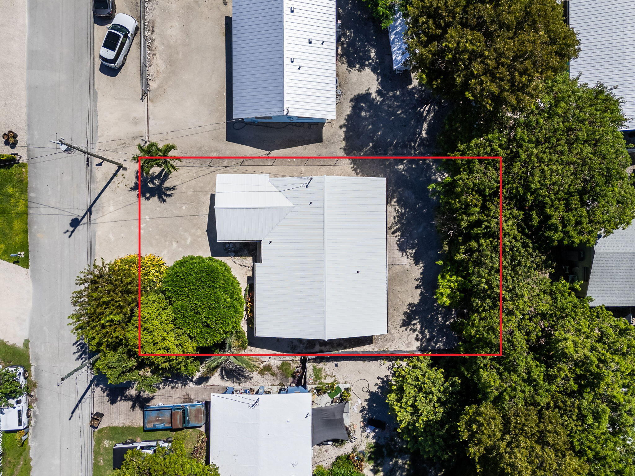 Aerial view of a white-roofed home and red-outlined lot surrounded by trees beside a road.