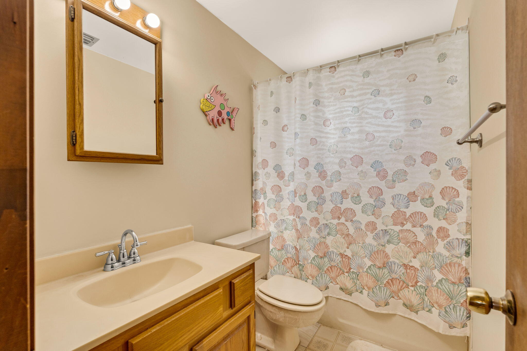 Bathroom with a wood vanity and mirror, toilet, and colorful seashell-patterned shower curtain.