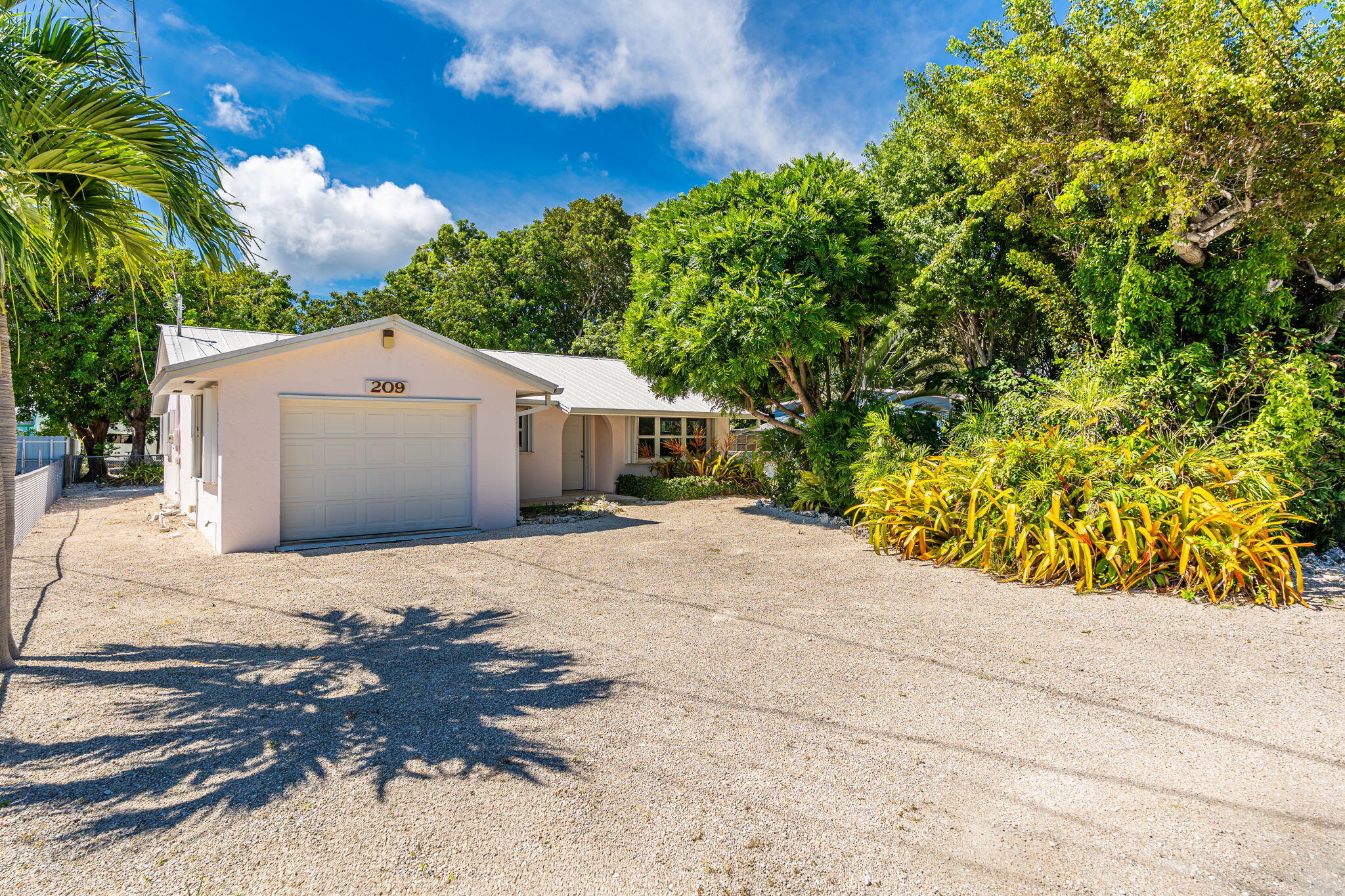 Single-story home with attached garage, gravel driveway, and dense tropical landscaping.