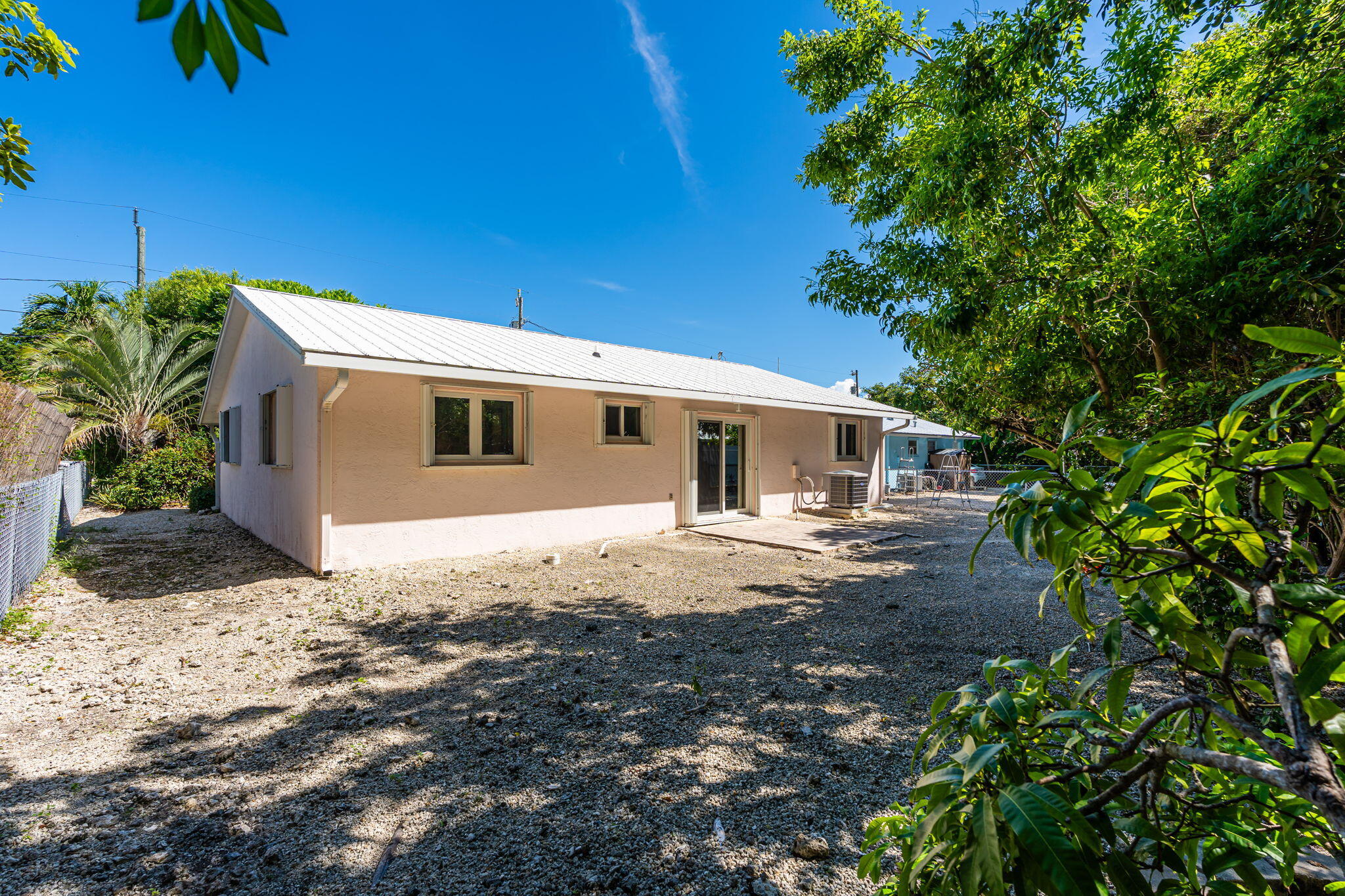 Single-story home with a white metal roof, sliding glass door, and tree-shaded gravel yard.