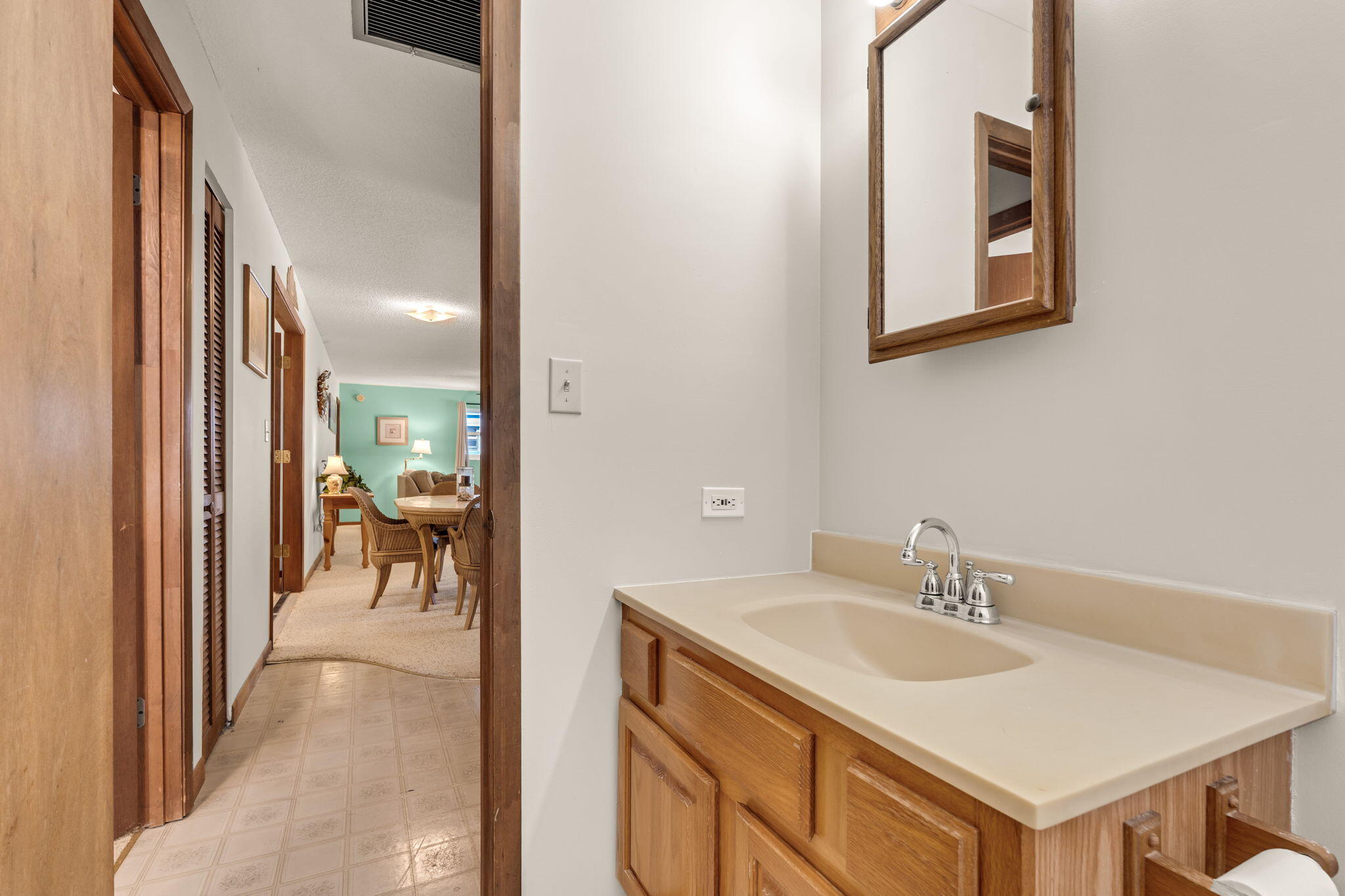 Bathroom vanity with a beige sink counter and wood cabinets beside a hallway opening to a furnished living area.