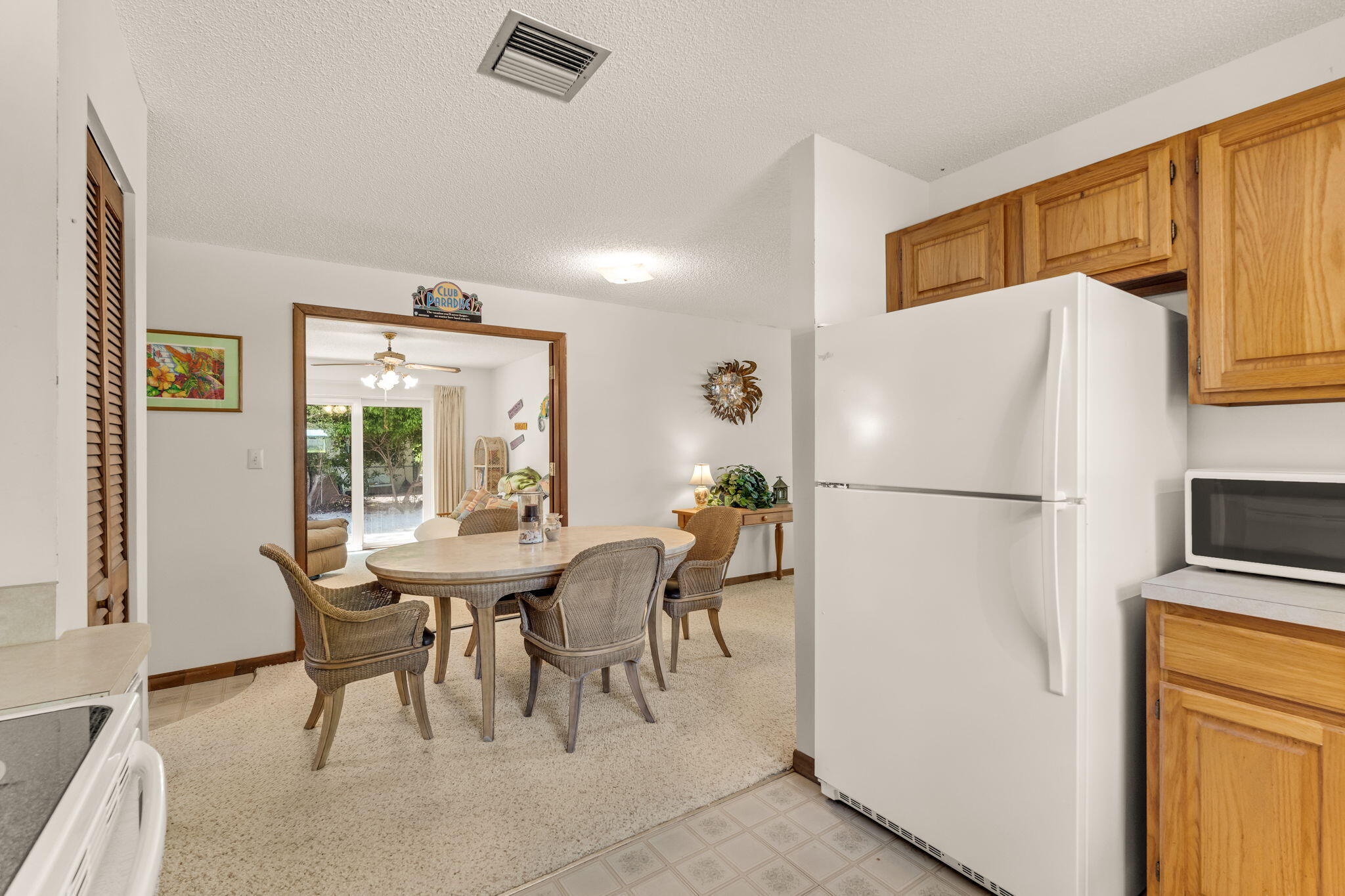 Dining area with a round table and wicker chairs beside a kitchen with wood cabinets and a white refrigerator.