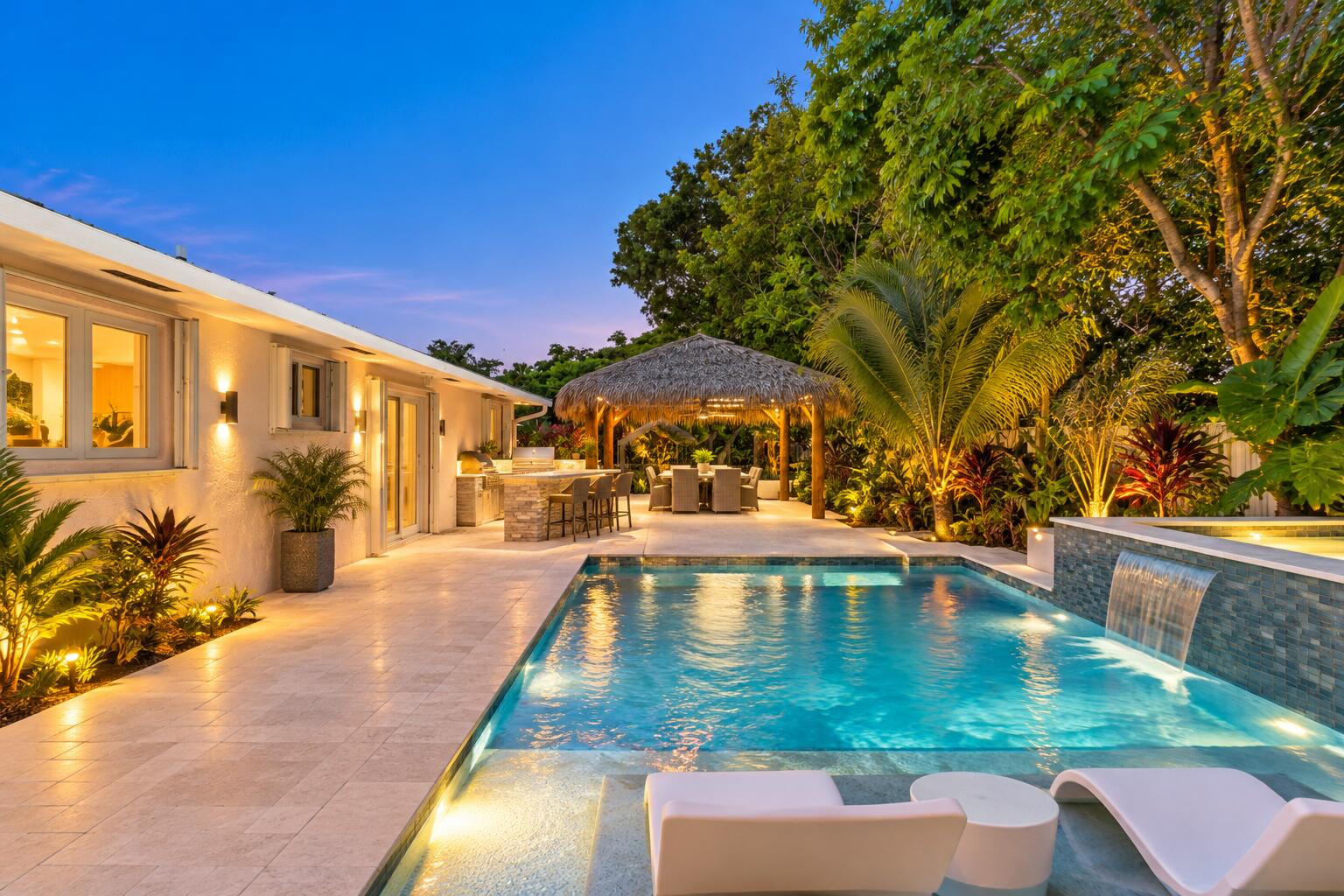 Backyard pool and patio at dusk, with a thatched pavilion, stone waterfall, and tropical landscaping.