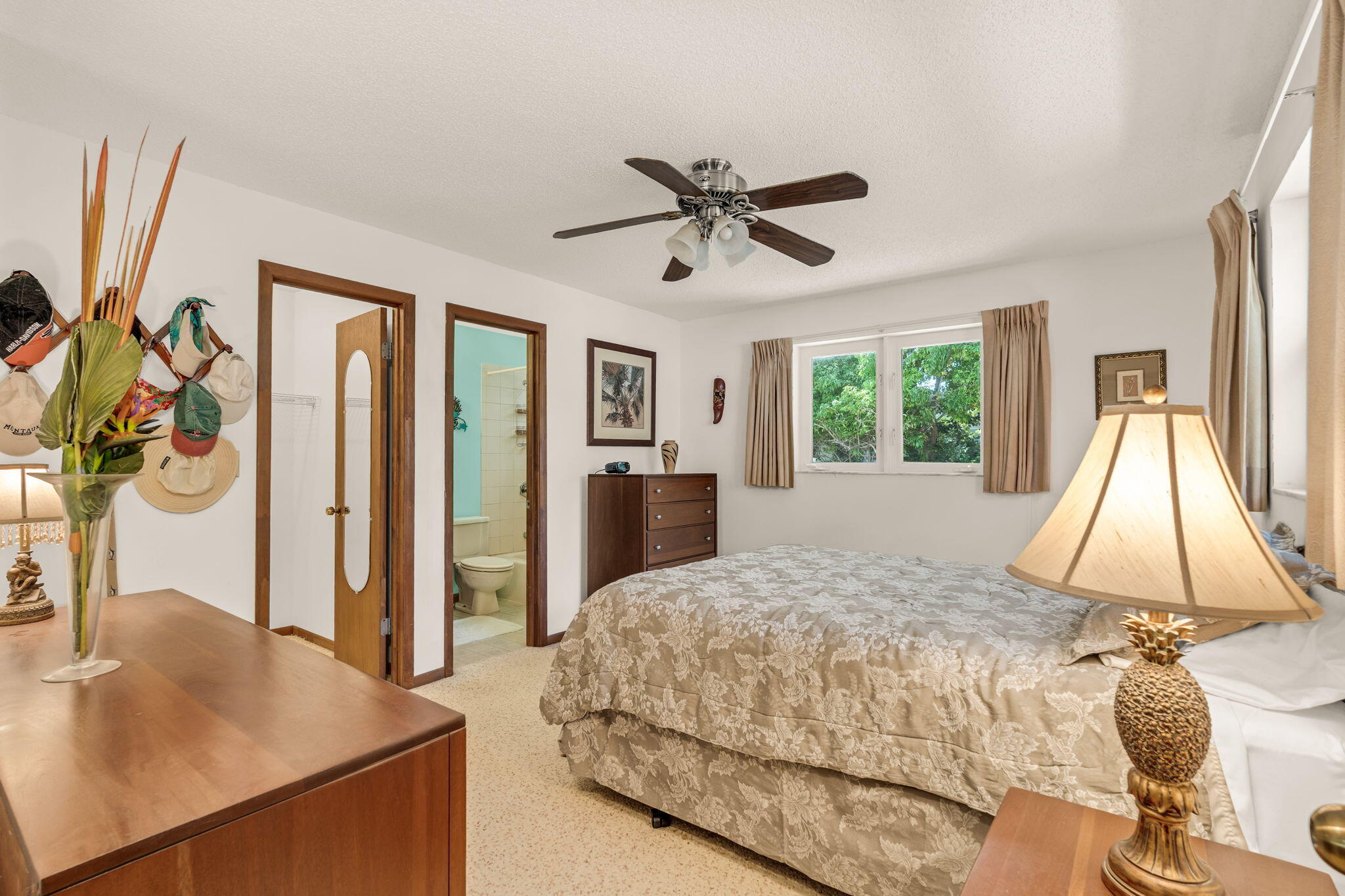 Furnished bedroom with a floral quilted bed, ceiling fan, and open doorway to a bathroom.