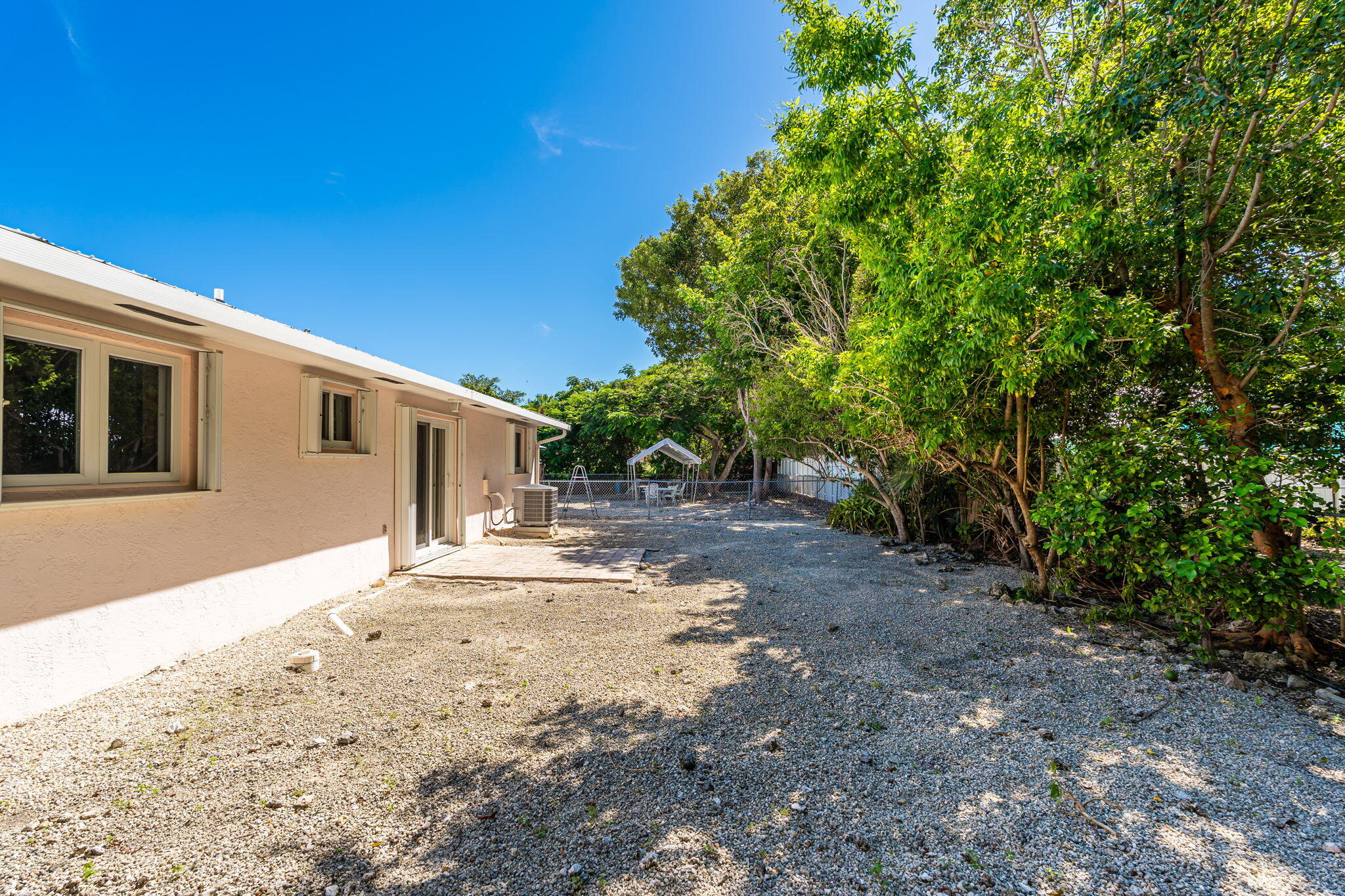 Gravel side yard beside a beige home, shaded by mature trees and enclosed by a chain-link fence.