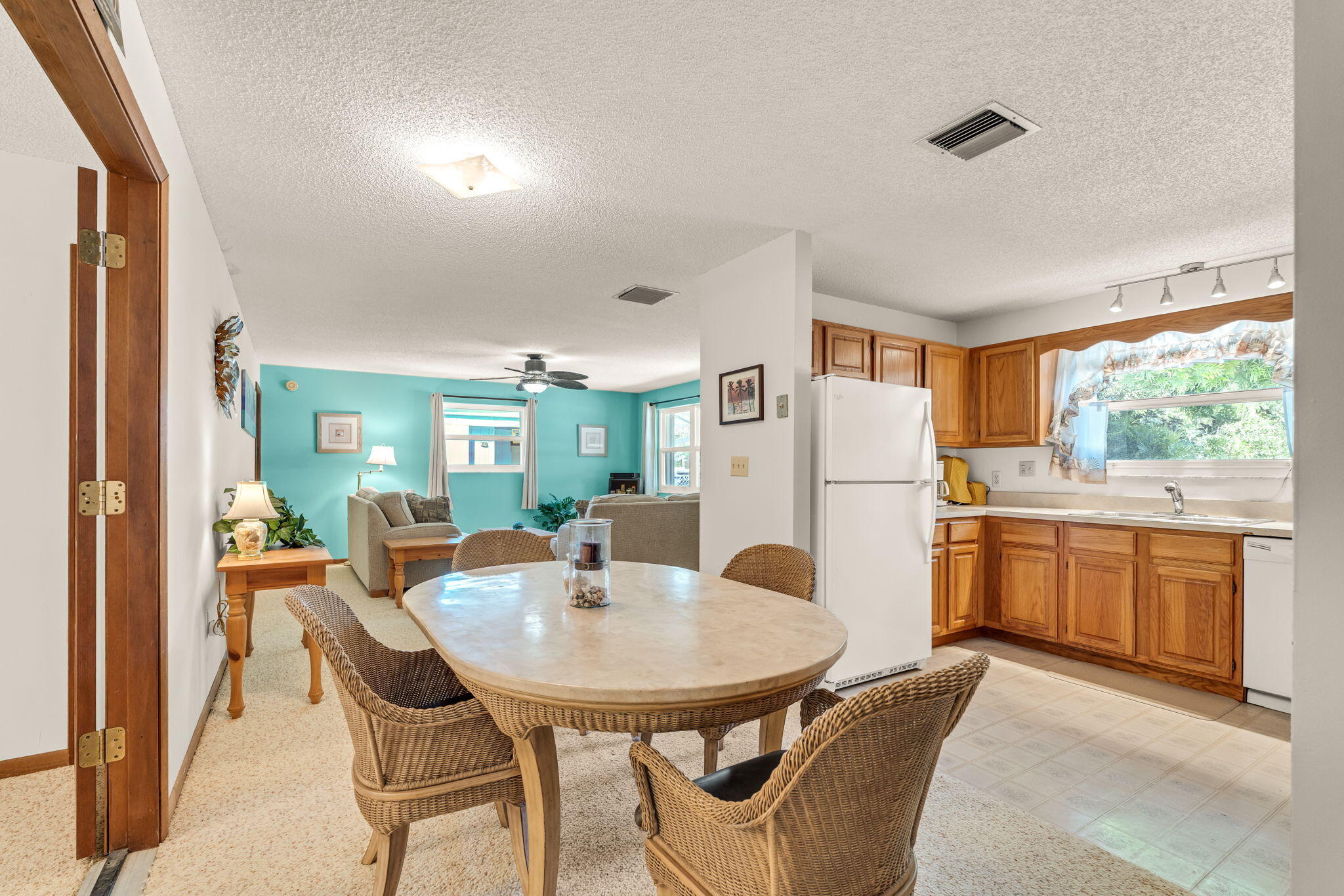 Open-plan kitchen and dining area with wood cabinets, wicker chairs, and a turquoise living room beyond.