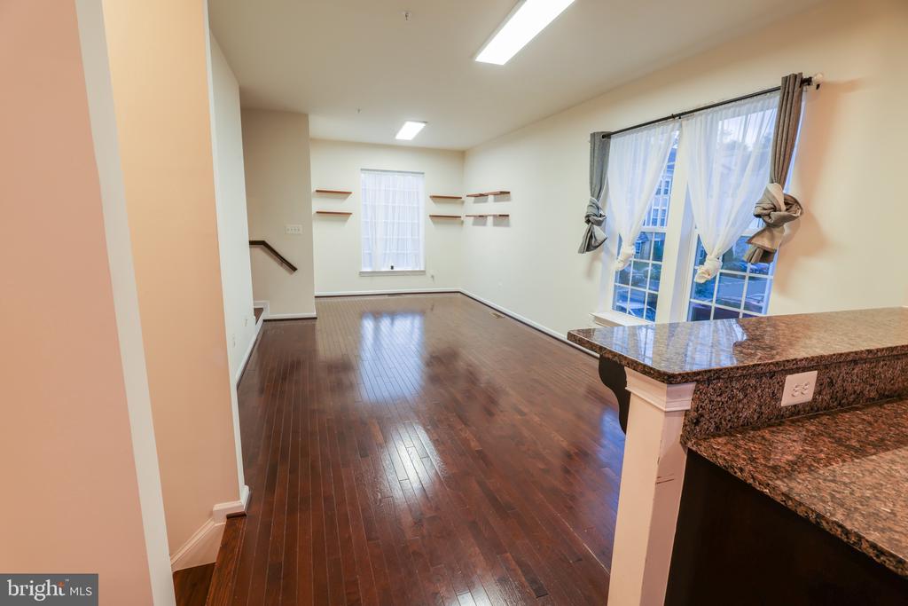 Empty room with polished hardwood floors, curtained windows, wall shelves, and a granite kitchen counter.