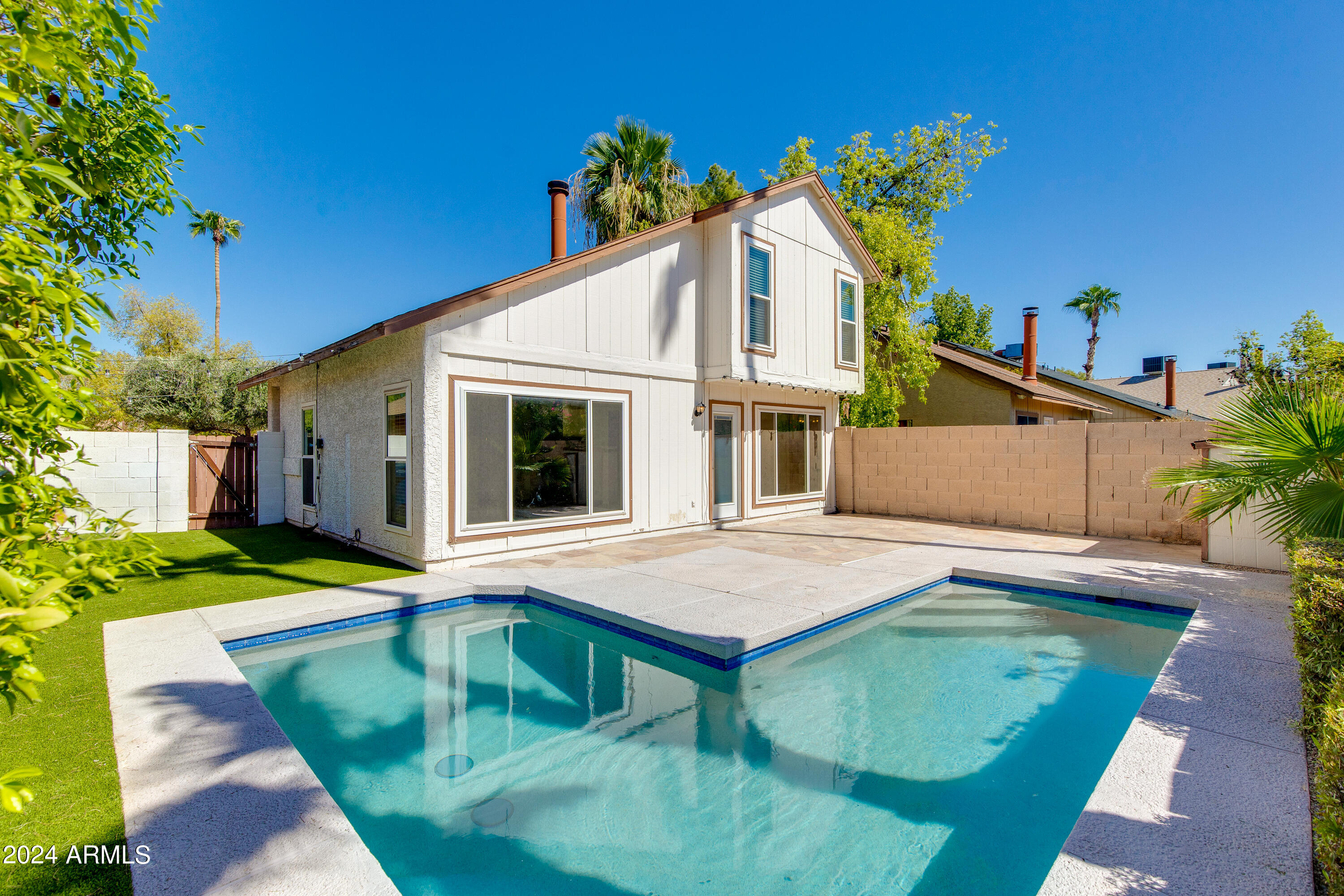 Backyard pool beside a two-story white home, with a paved deck, artificial turf, and block-wall fencing.