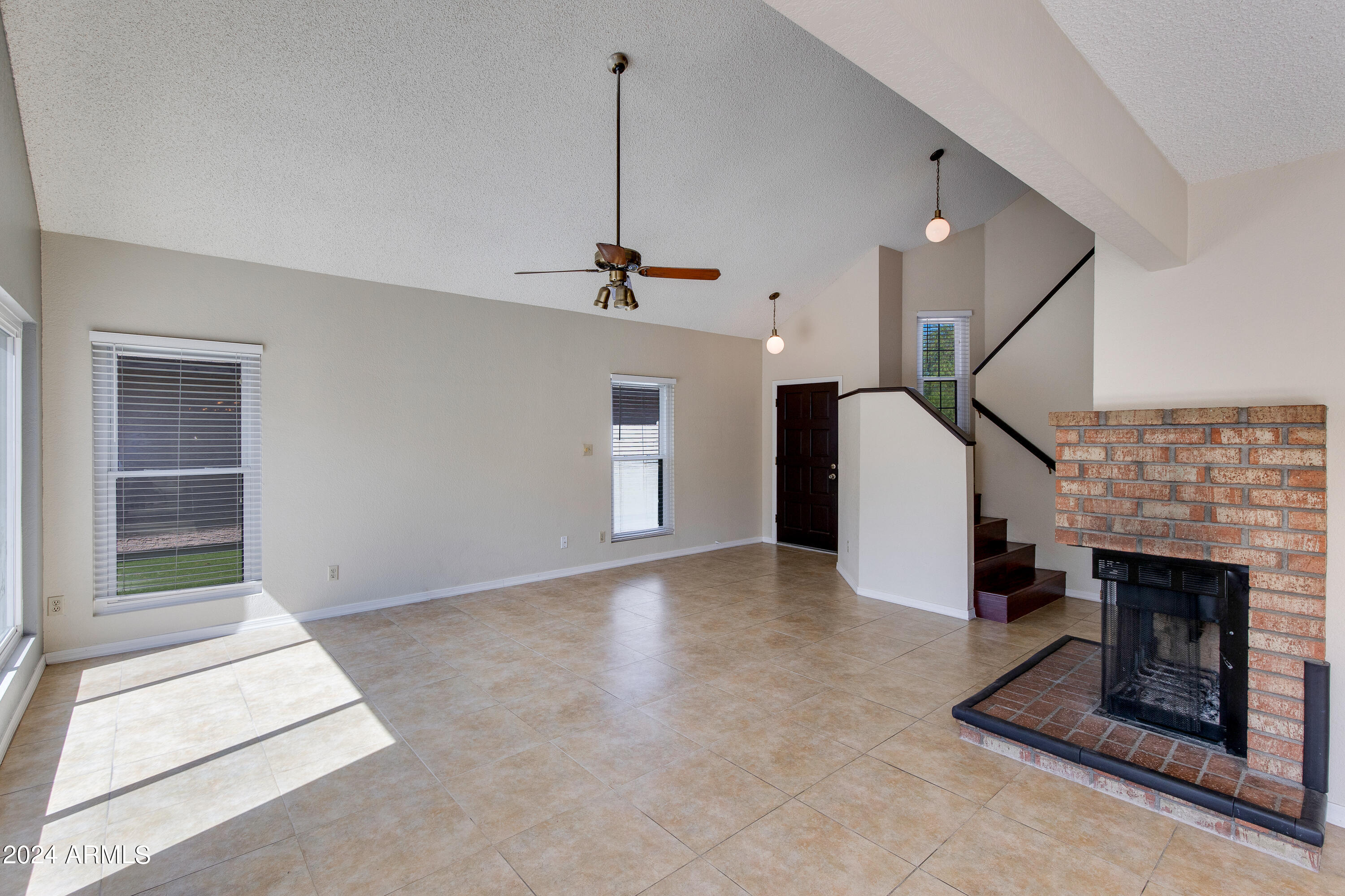 Vacant living room with a brick fireplace, vaulted ceiling, and staircase.