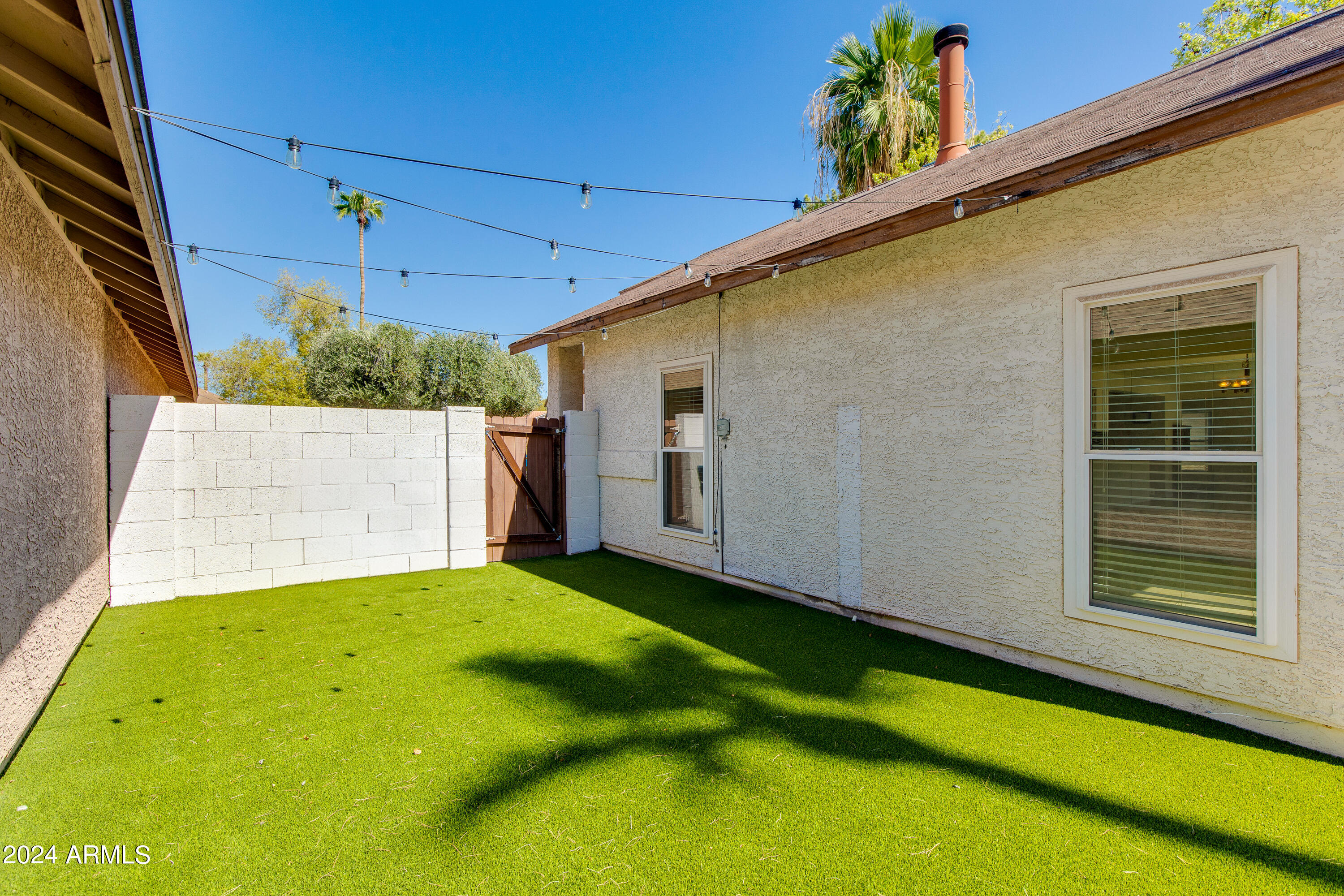 Enclosed backyard with artificial turf, overhead string lights, and a wooden gate beside the stucco house.