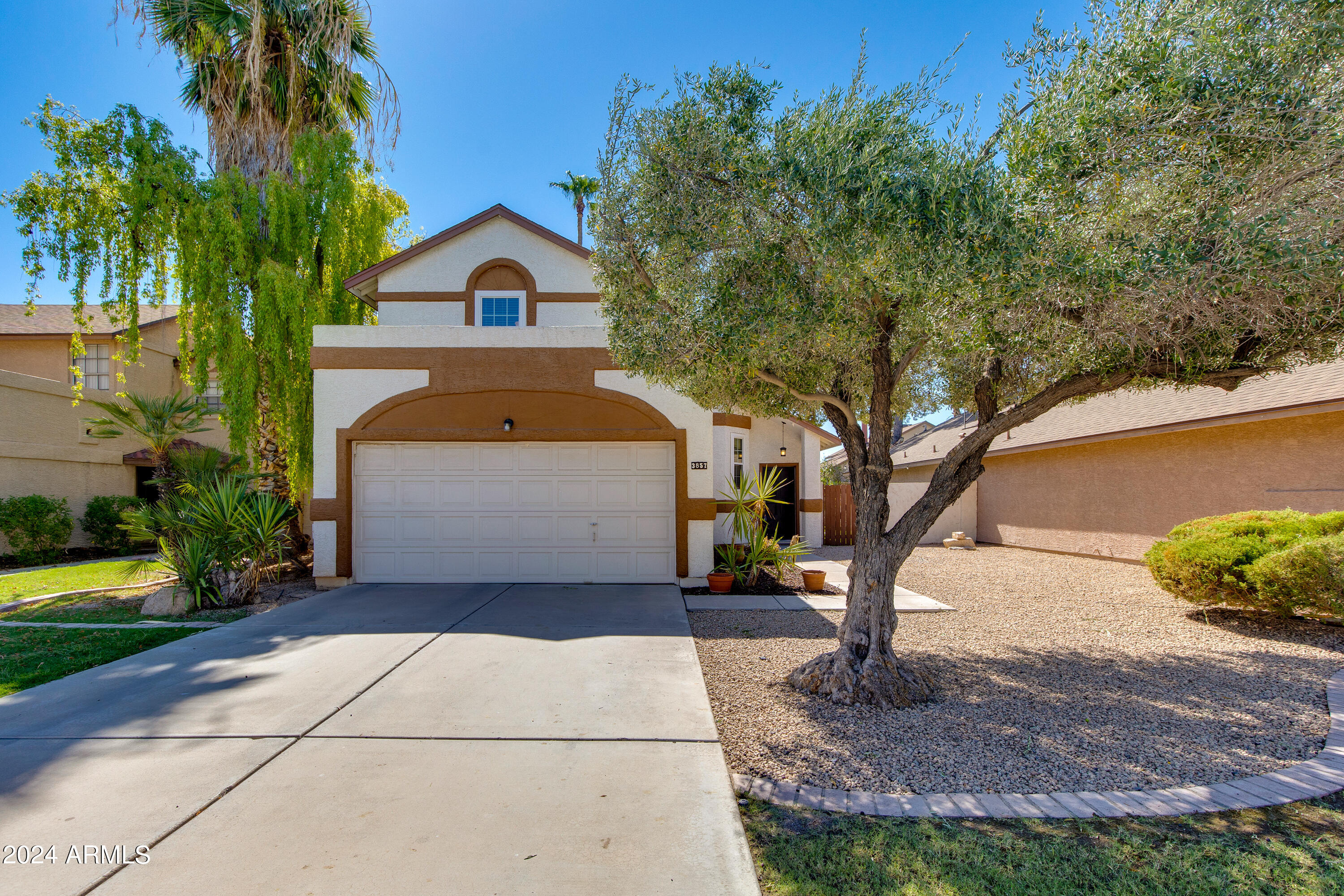 Two-story stucco home with a double garage, shaded driveway, and gravel front yard.