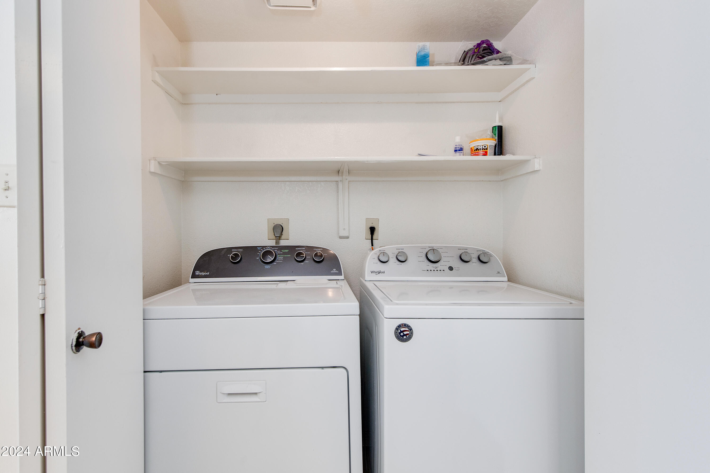 Laundry room with a top-load washer and dryer beneath two storage shelves.