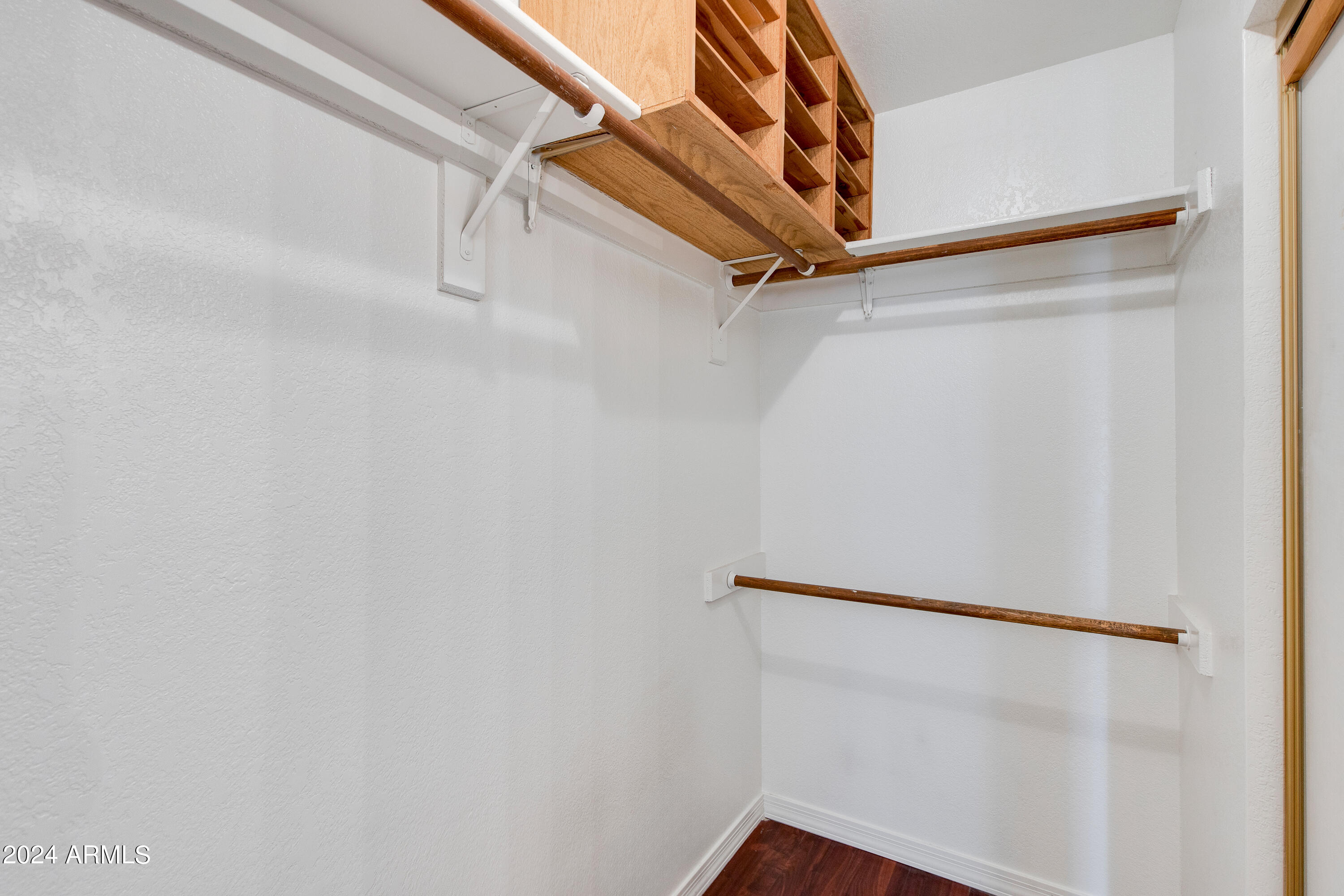 Walk-in closet with overhead wooden shelves, hanging rods, and dark wood flooring.