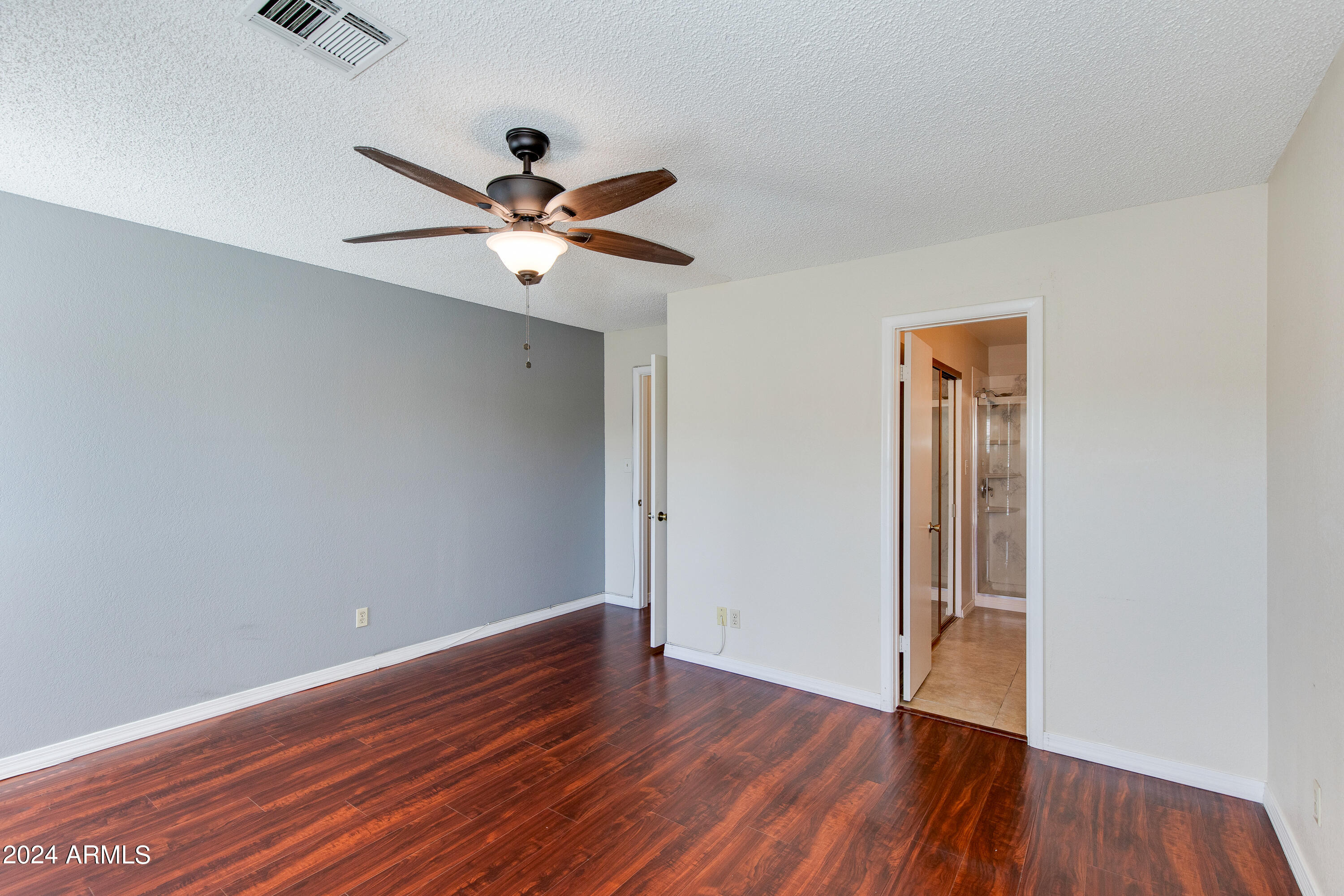 Empty bedroom with dark wood flooring and a ceiling fan, opening to a bathroom with a glass shower.