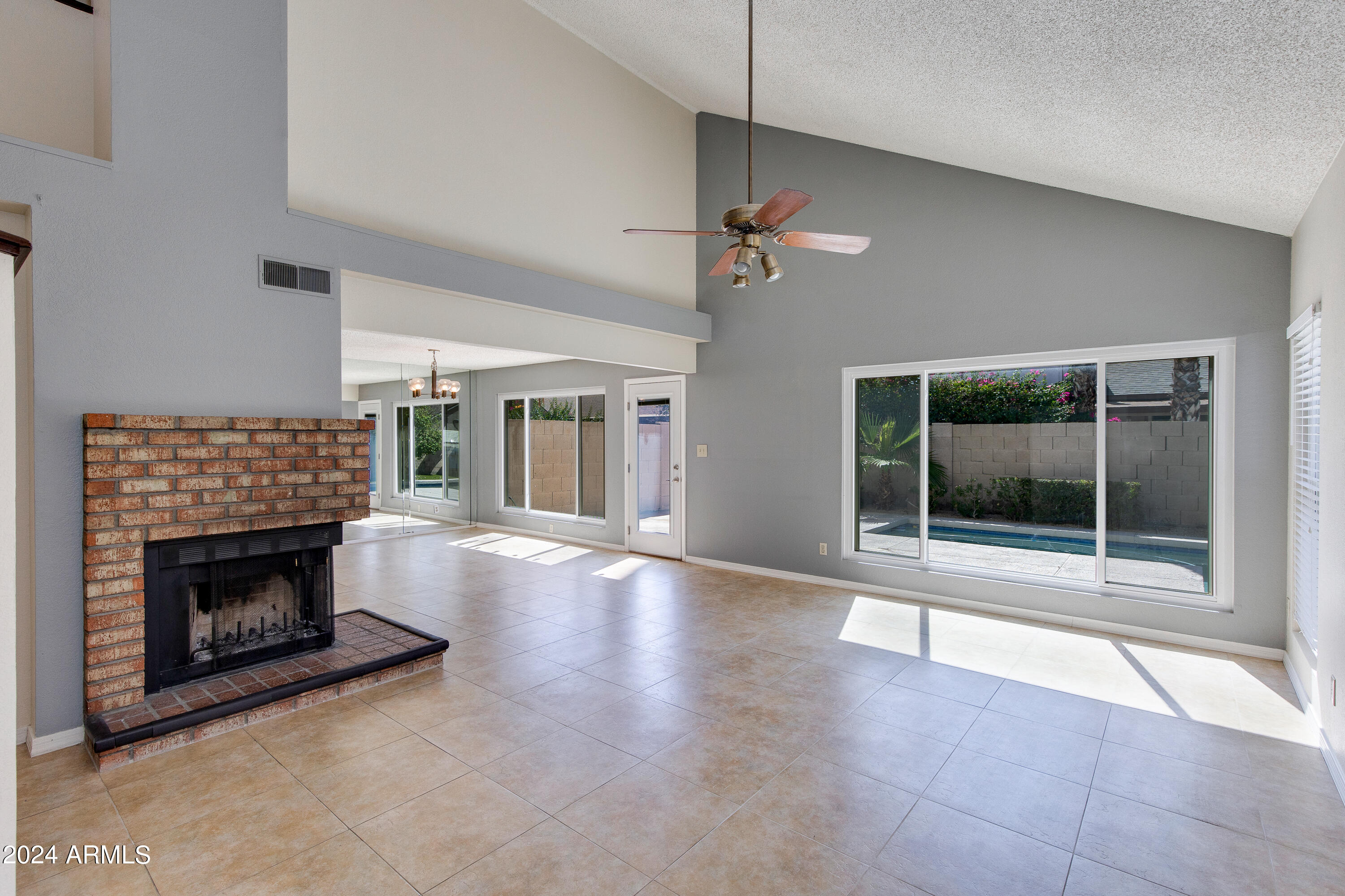 Living room with a brick fireplace, vaulted ceiling, and large windows overlooking a pool.