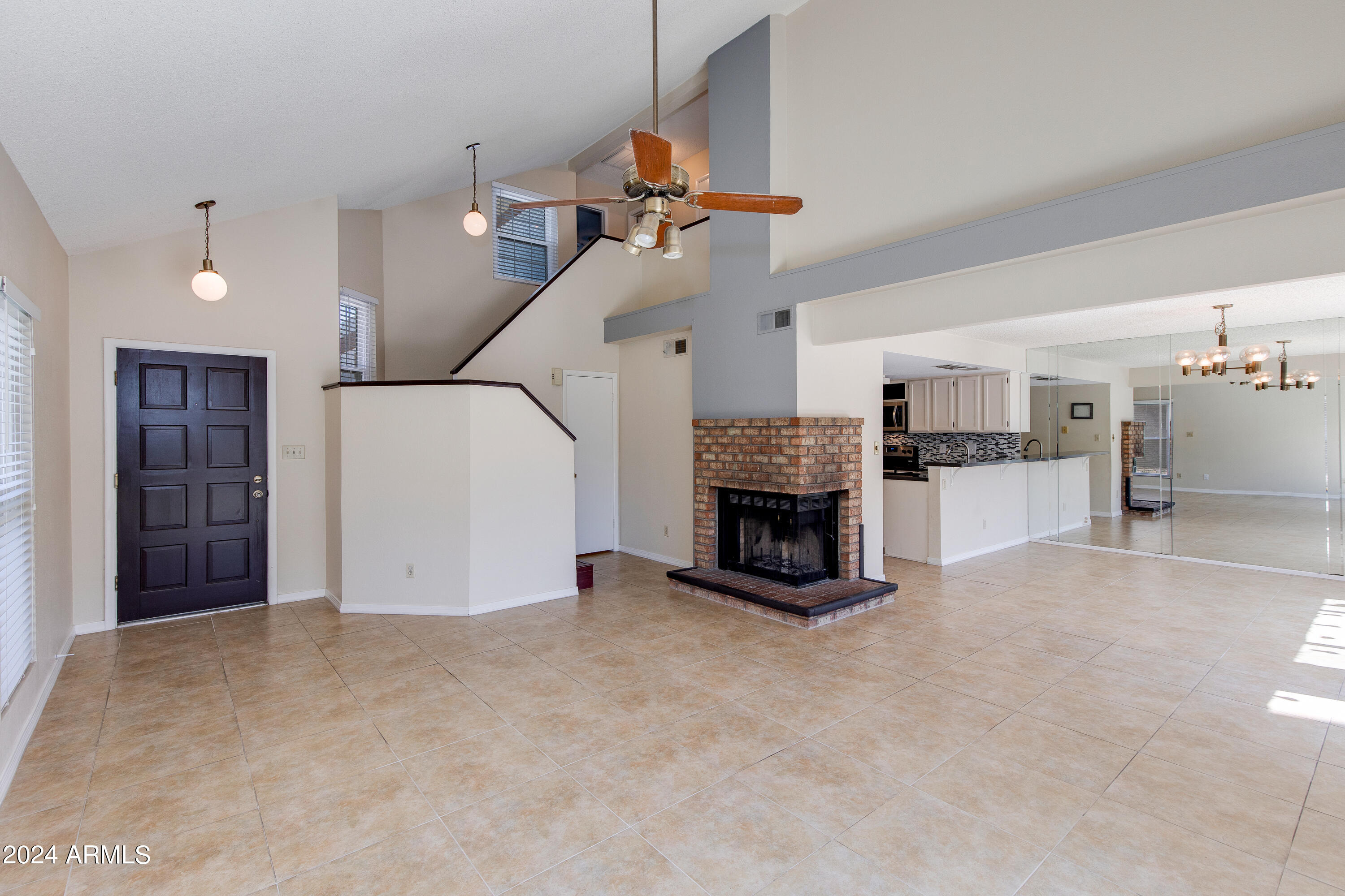 Open living room with brick fireplace, tile floors, staircase, and adjoining kitchen.