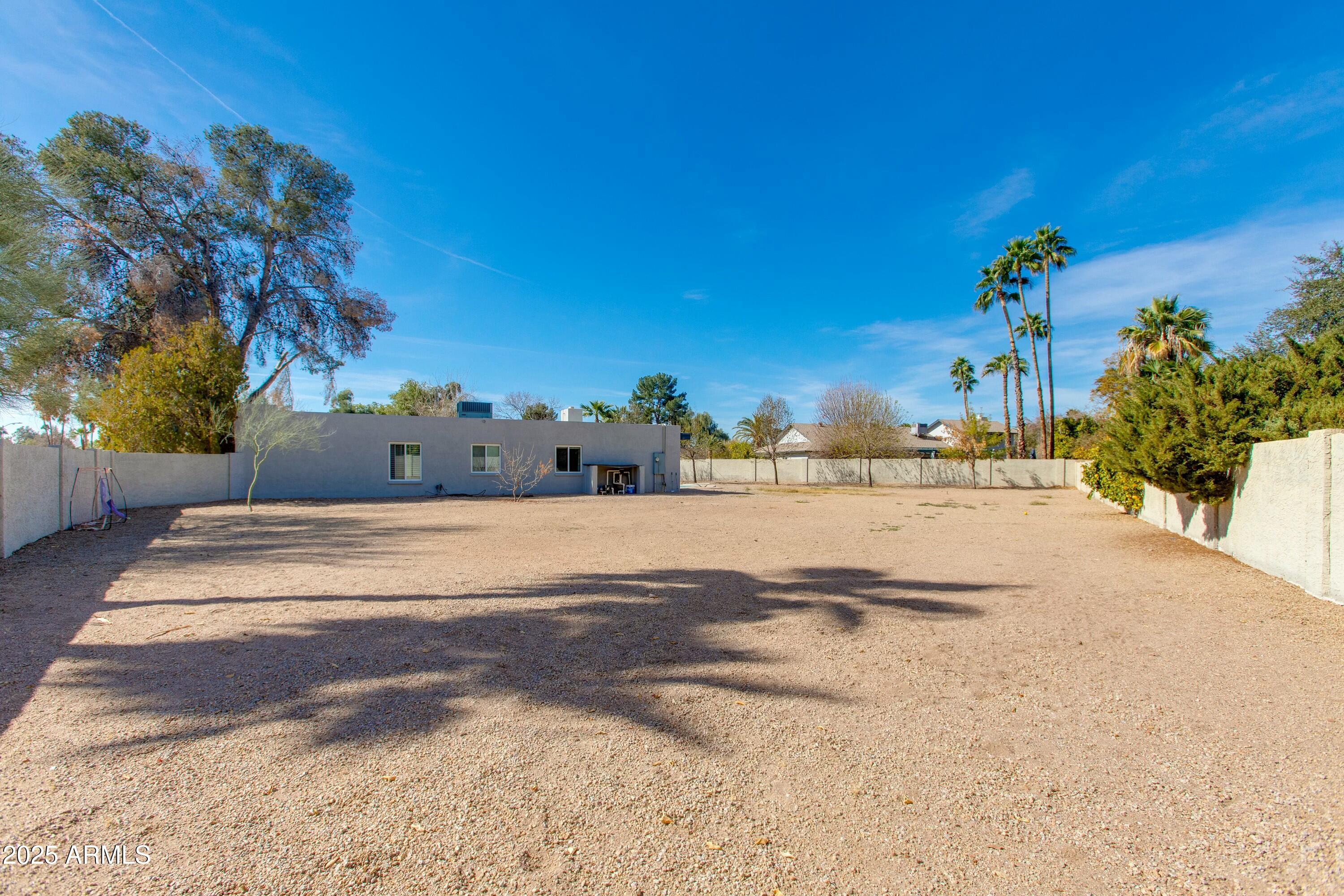 Gravel backyard enclosed by walls, with a low gray home and tall palm trees.