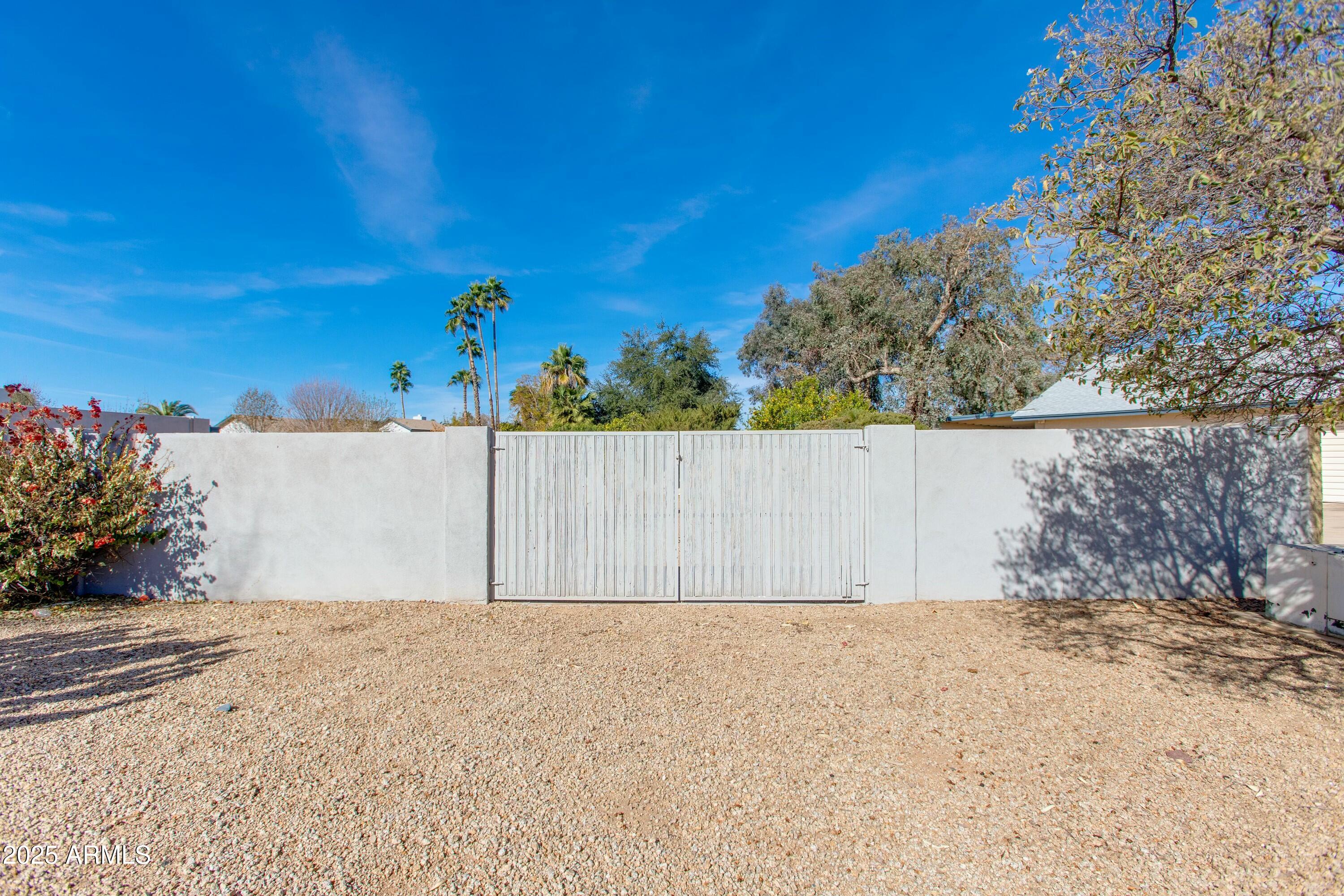 Gravel yard enclosed by a white wall with a wide double gate and palm trees beyond.
