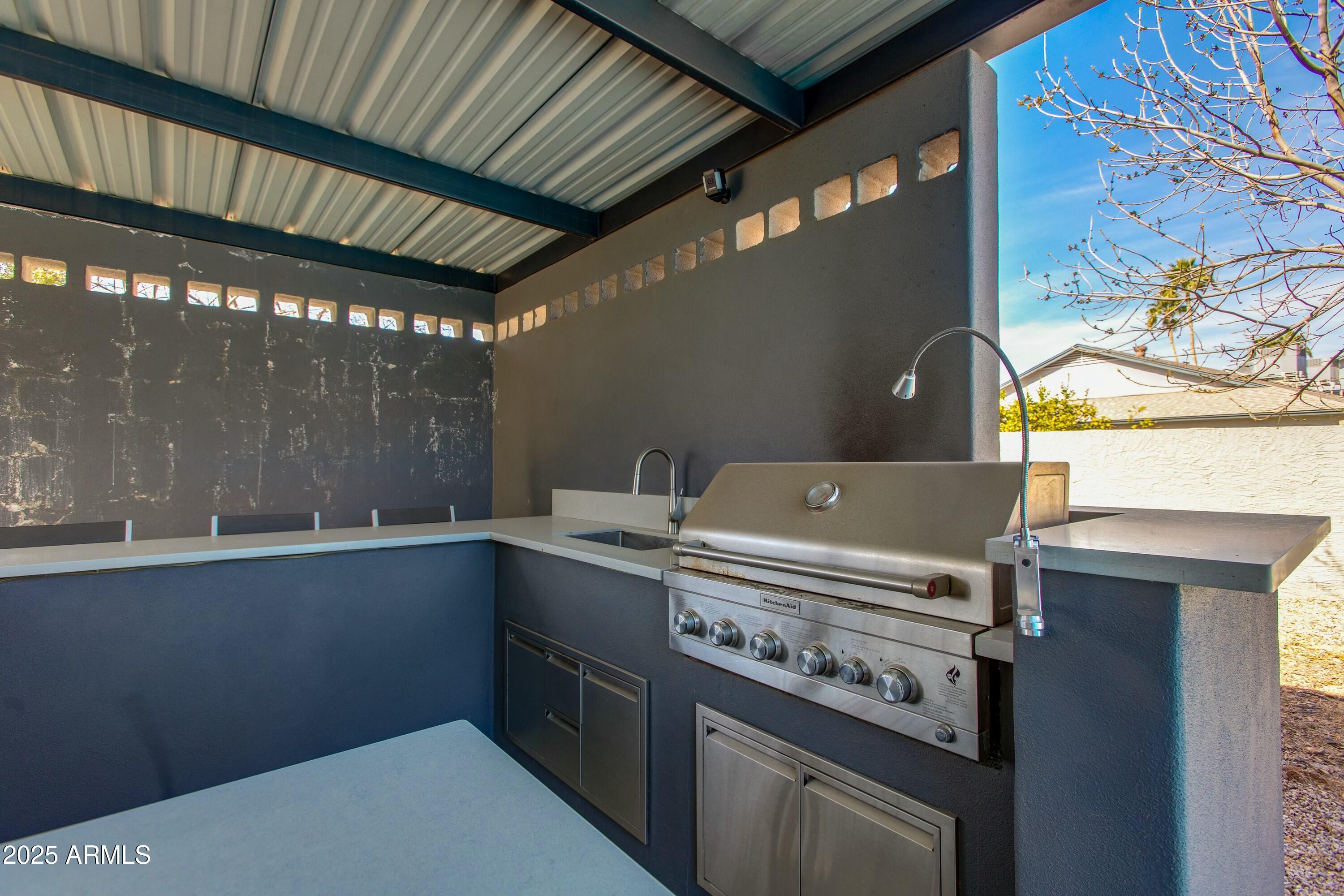 Covered outdoor kitchen with a built-in stainless steel grill, sink, and dark stone counters.