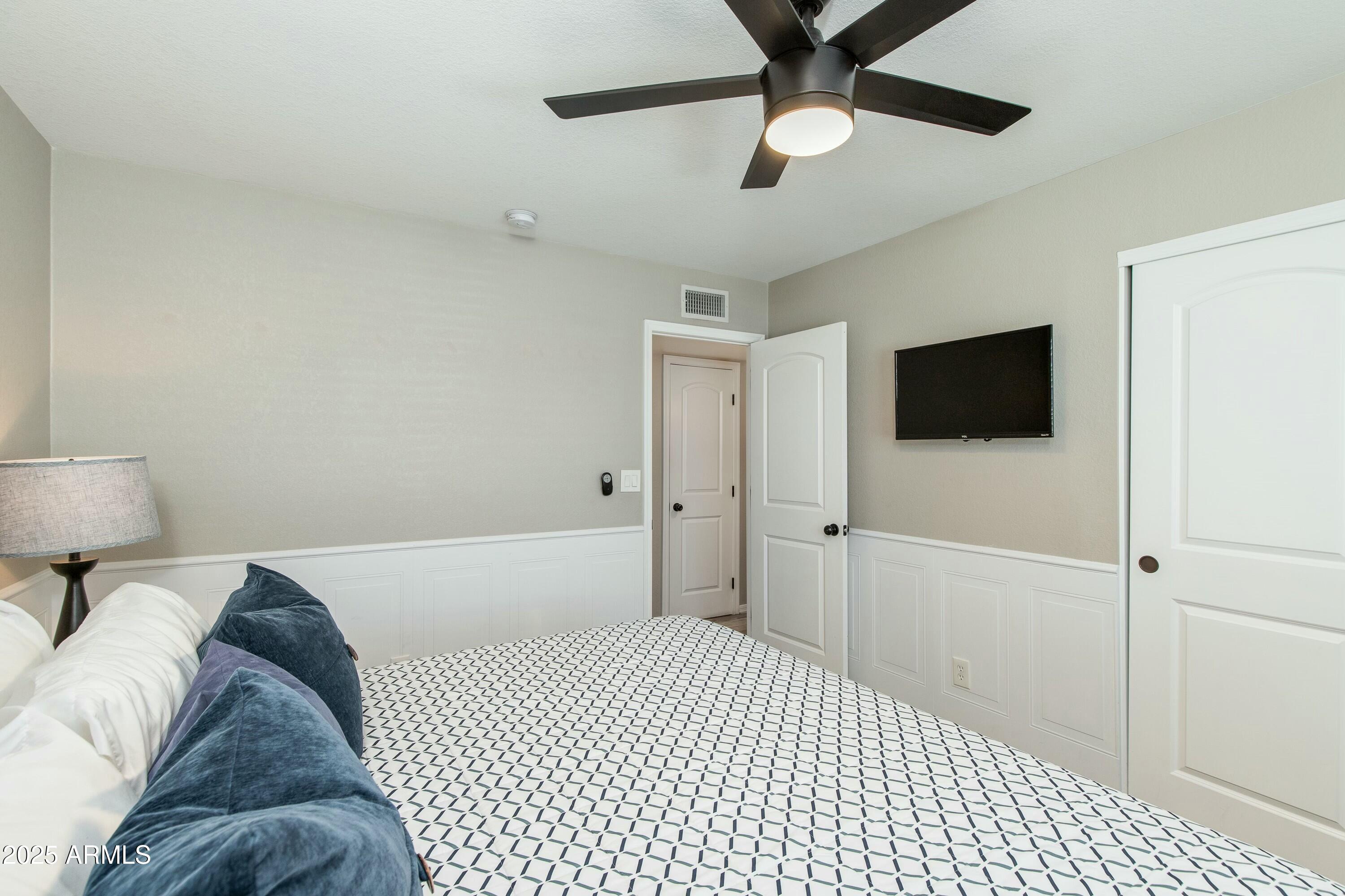 Bedroom with a geometric bedspread, blue pillows, wall-mounted TV, and ceiling fan.