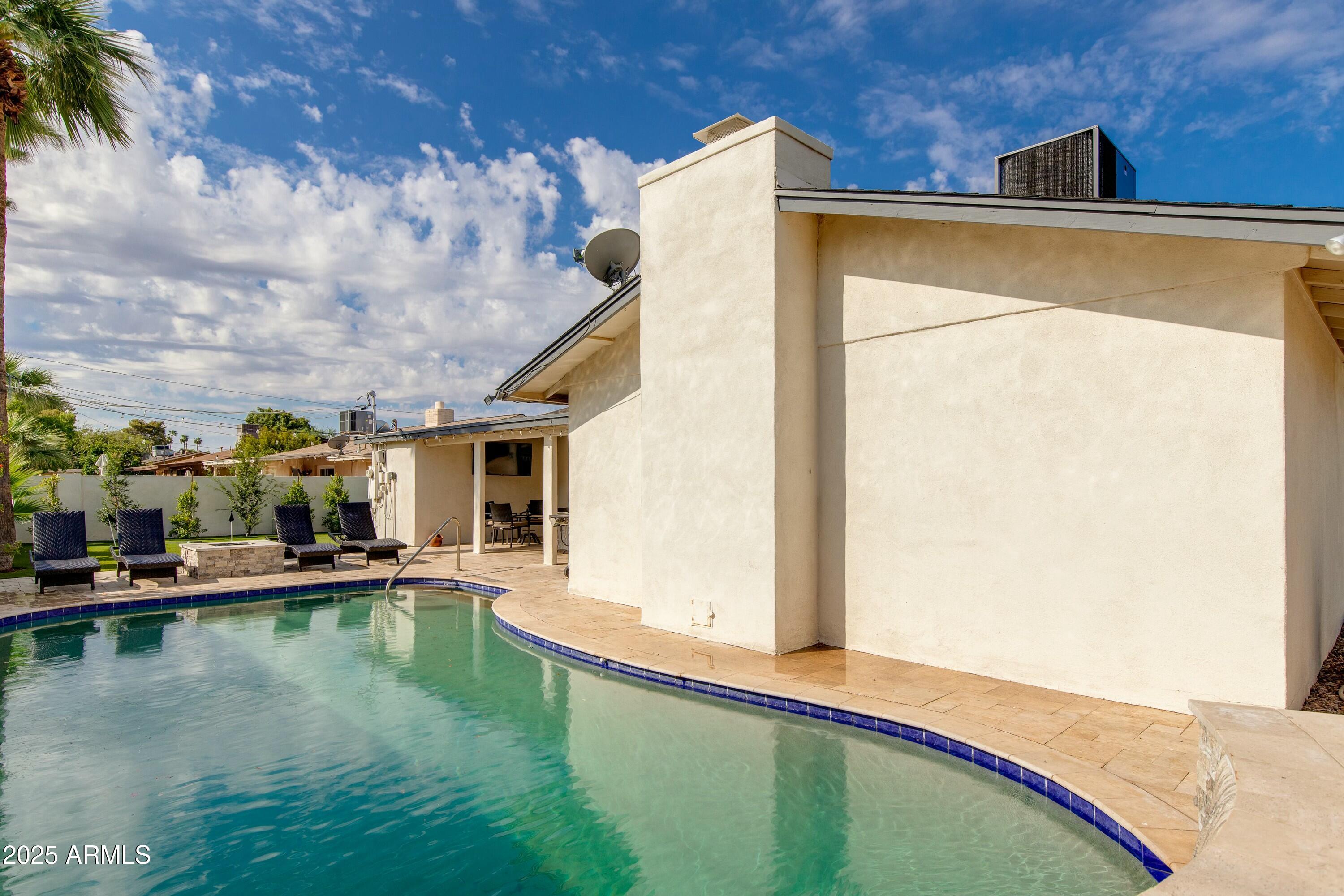 Curved backyard pool beside a stucco home, with a tiled patio and dark lounge chairs.