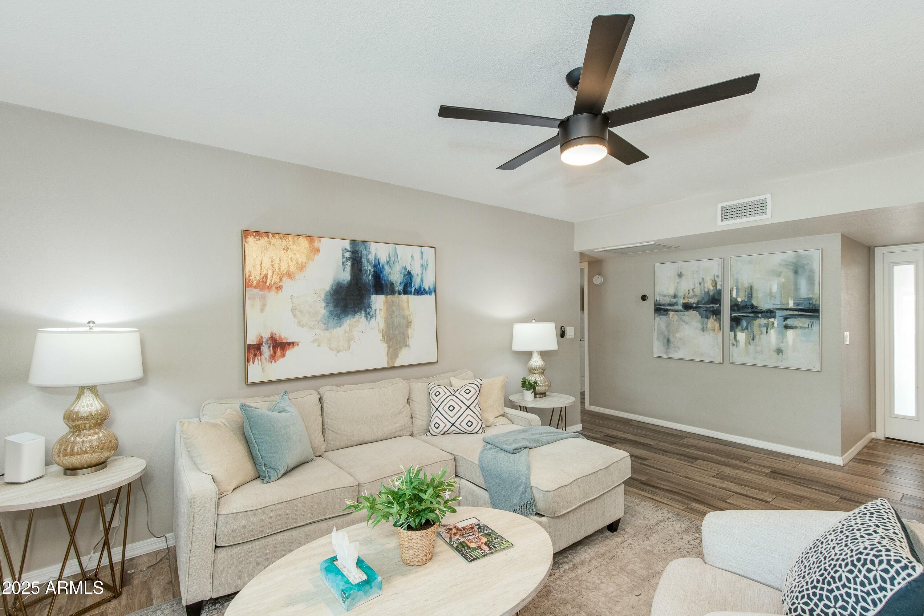 Staged living room with a beige sectional, abstract art, and dark ceiling fan.