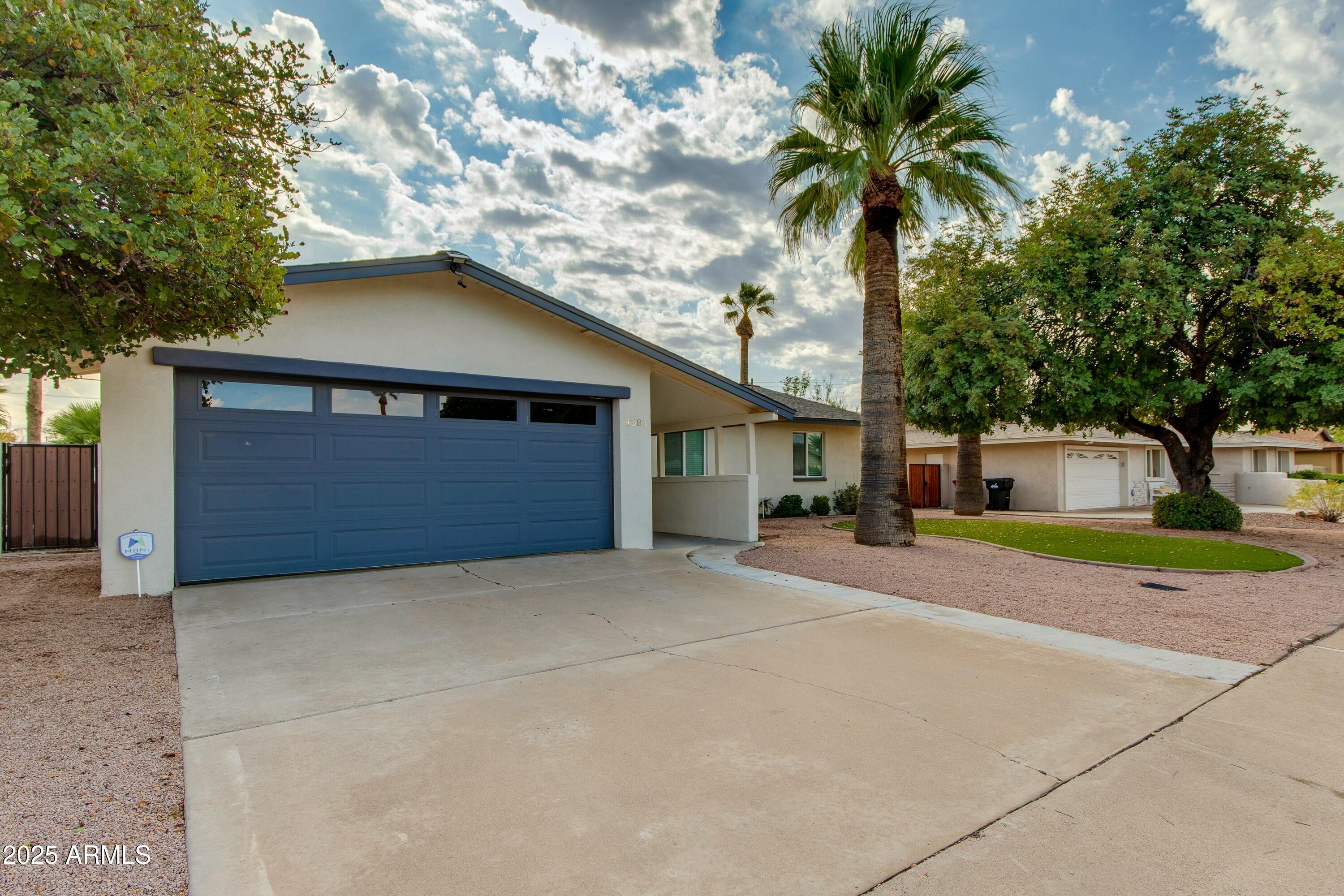 Single-story home with a blue double garage door, wide driveway, gravel yard, and palm trees.
