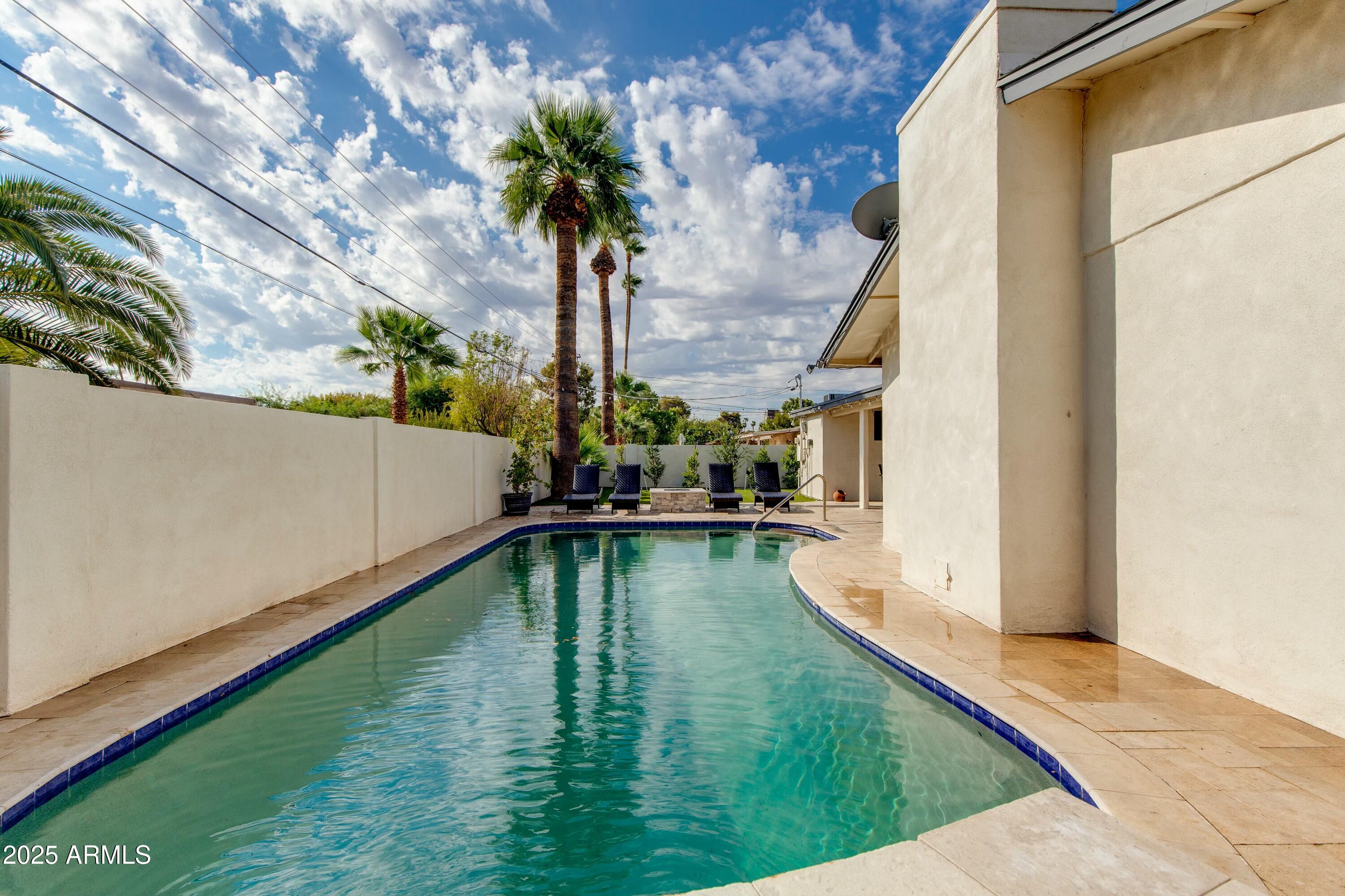Backyard pool beside a stucco home, with palm trees and lounge chairs along the far edge.