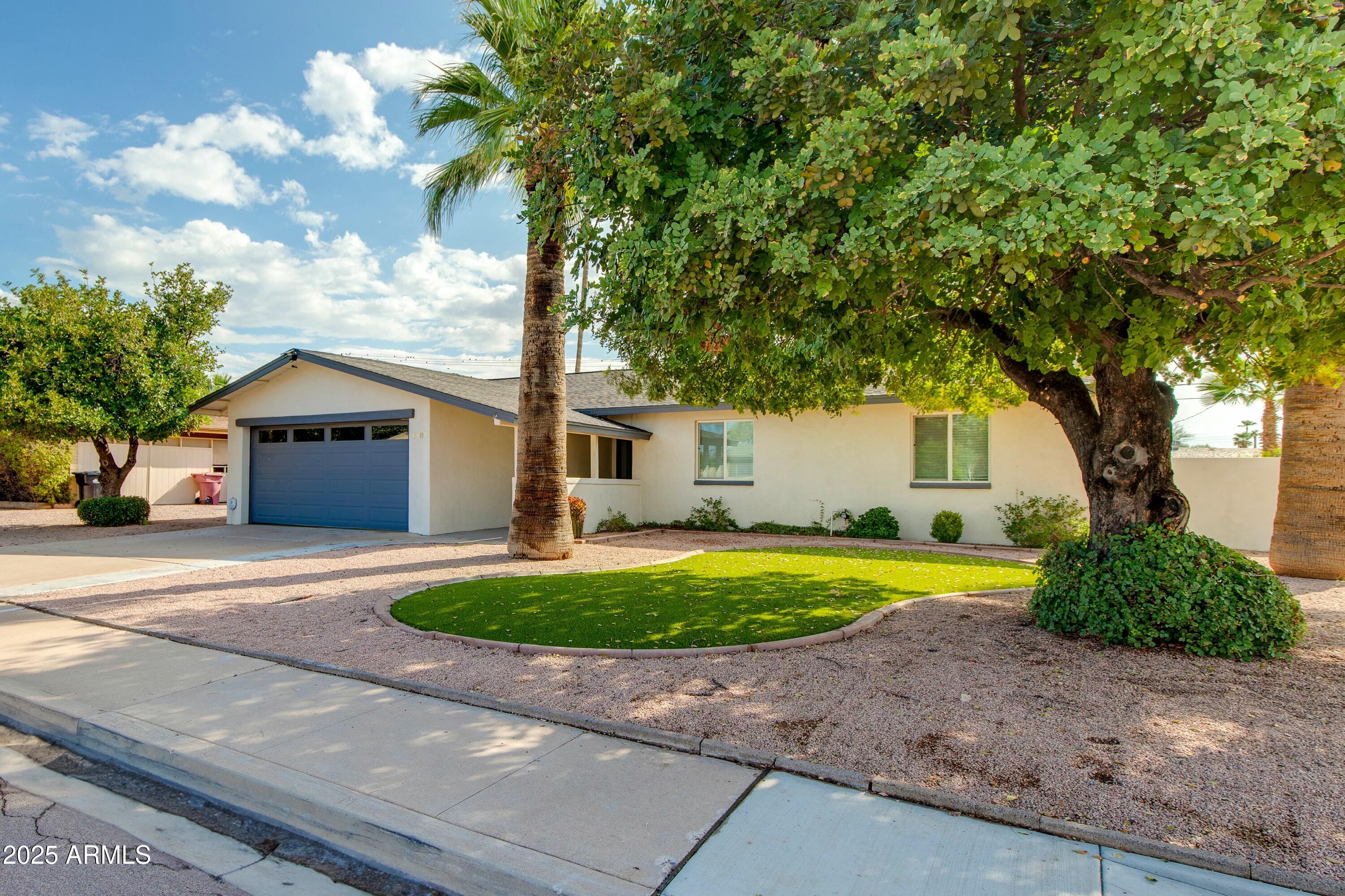 Single-story stucco home with a blue garage door, curved lawn, gravel yard, and mature shade trees.