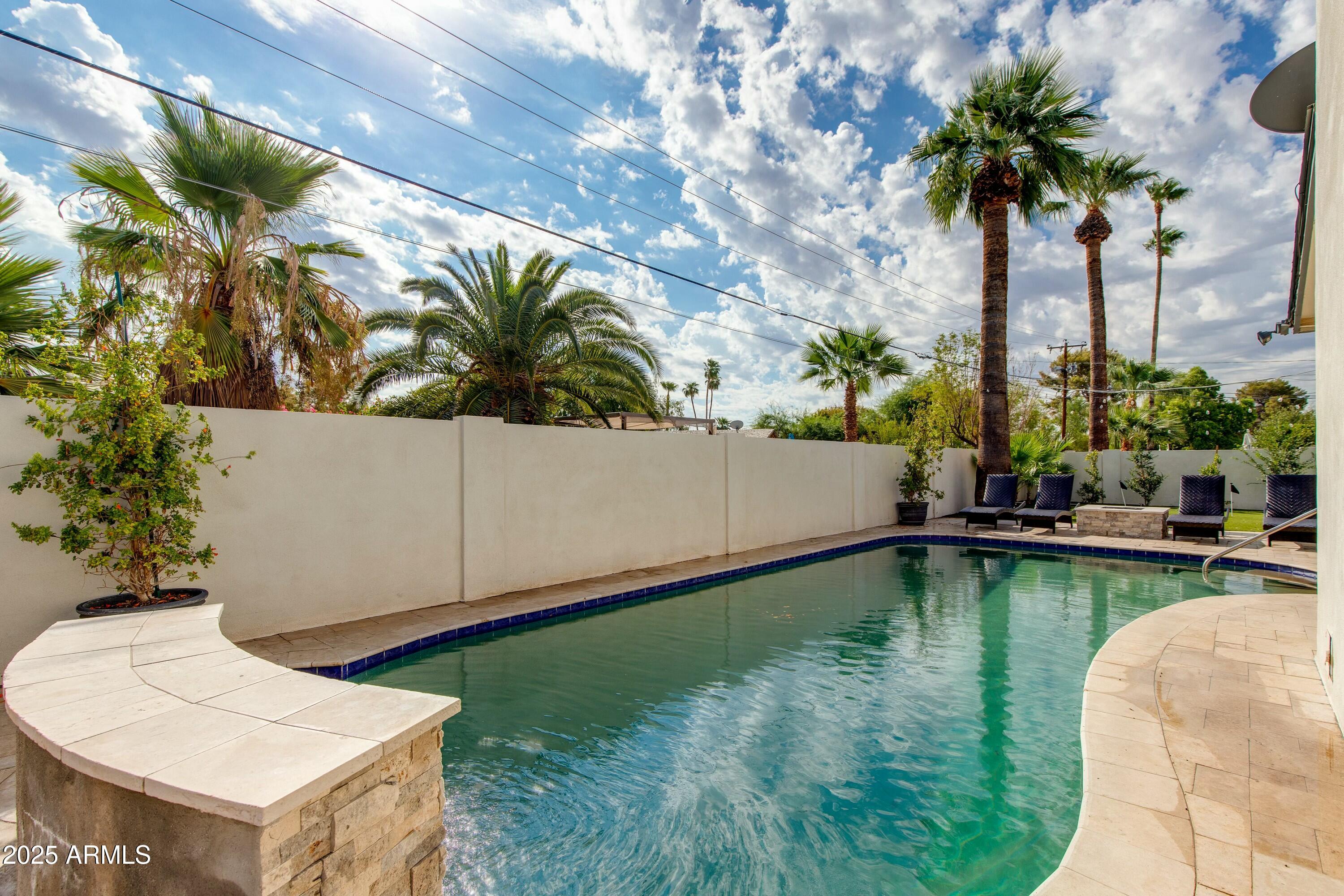 Backyard pool bordered by curved stone coping, palm trees, and patio seating.