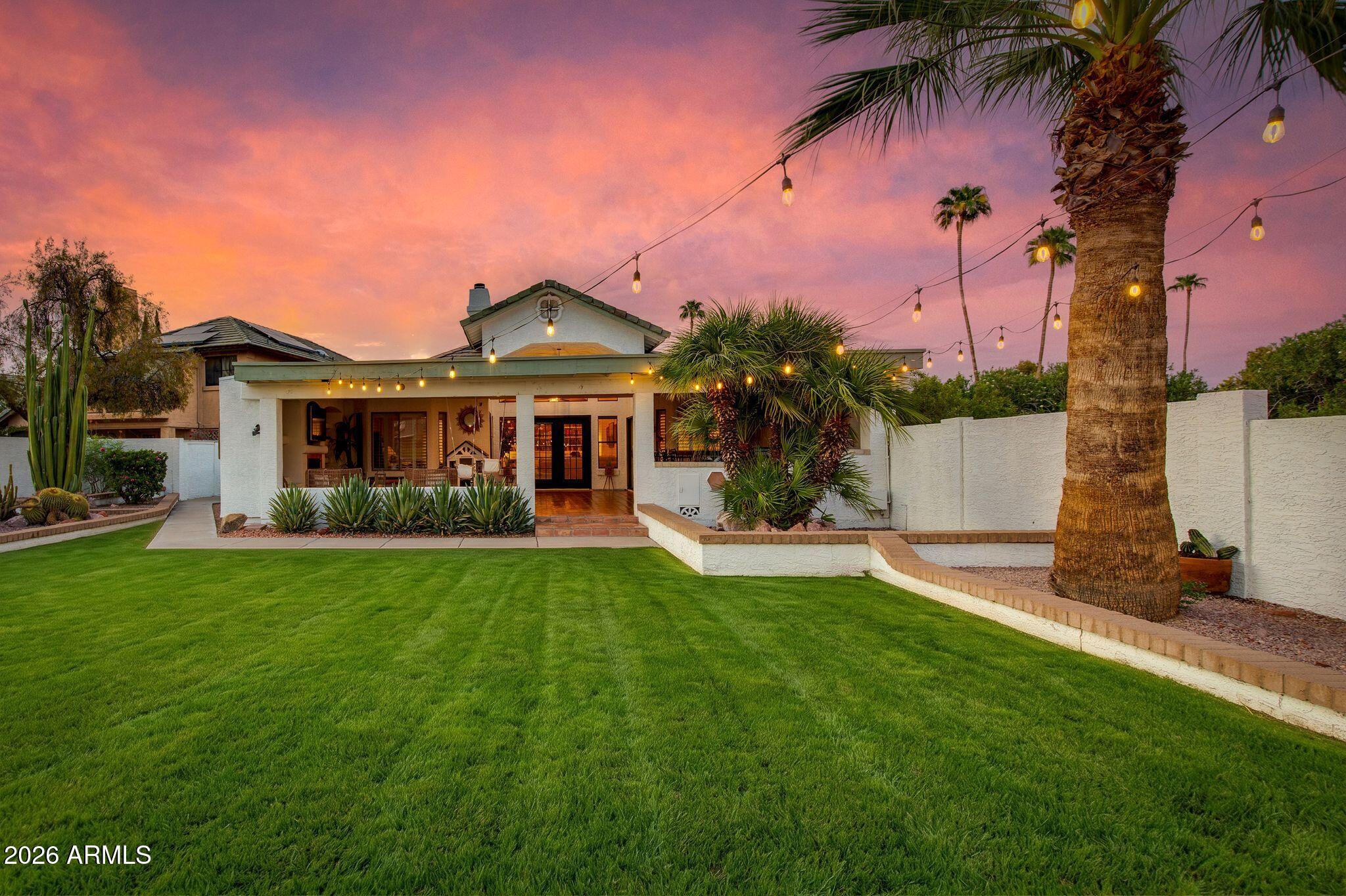 Home’s rear exterior with a covered patio, string lights, palms, and a broad lawn at dusk.