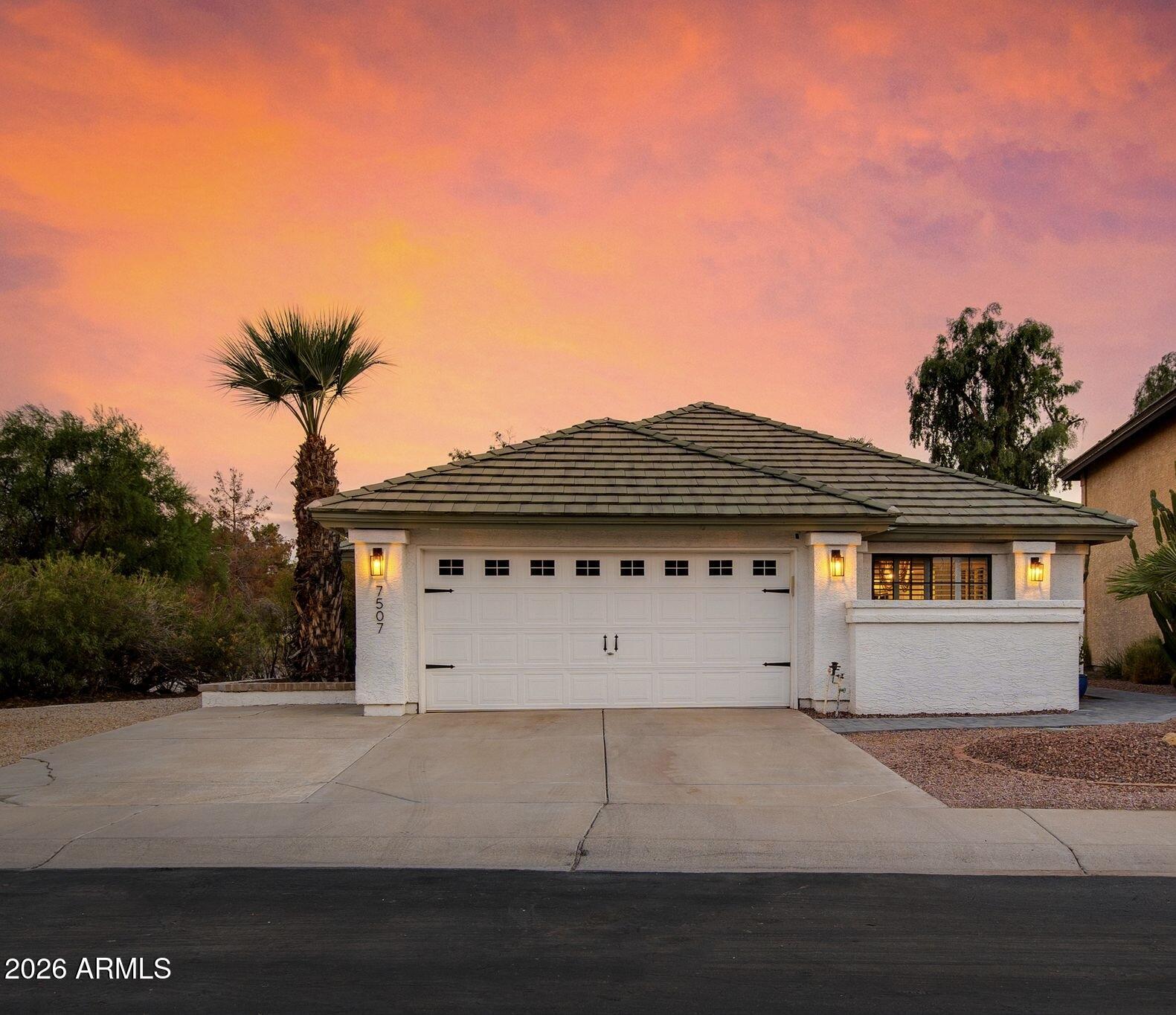Single-story stucco home with a double garage and tile roof beneath a vivid pink-orange dusk sky.